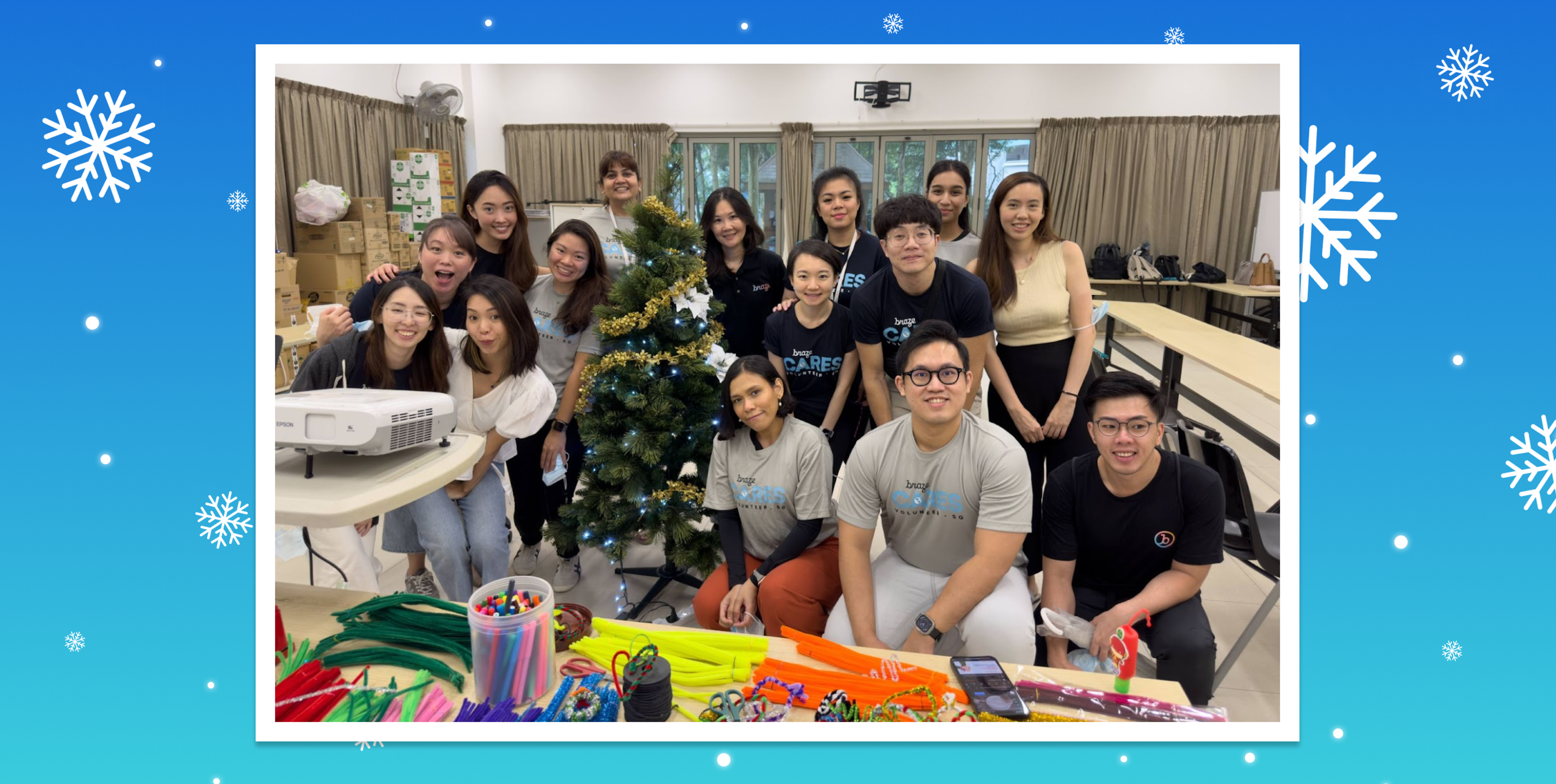 A group of people gathered around a decorated Christmas tree, smiling and posing for the photo. Colorful craft supplies are spread out on the table in front of them.