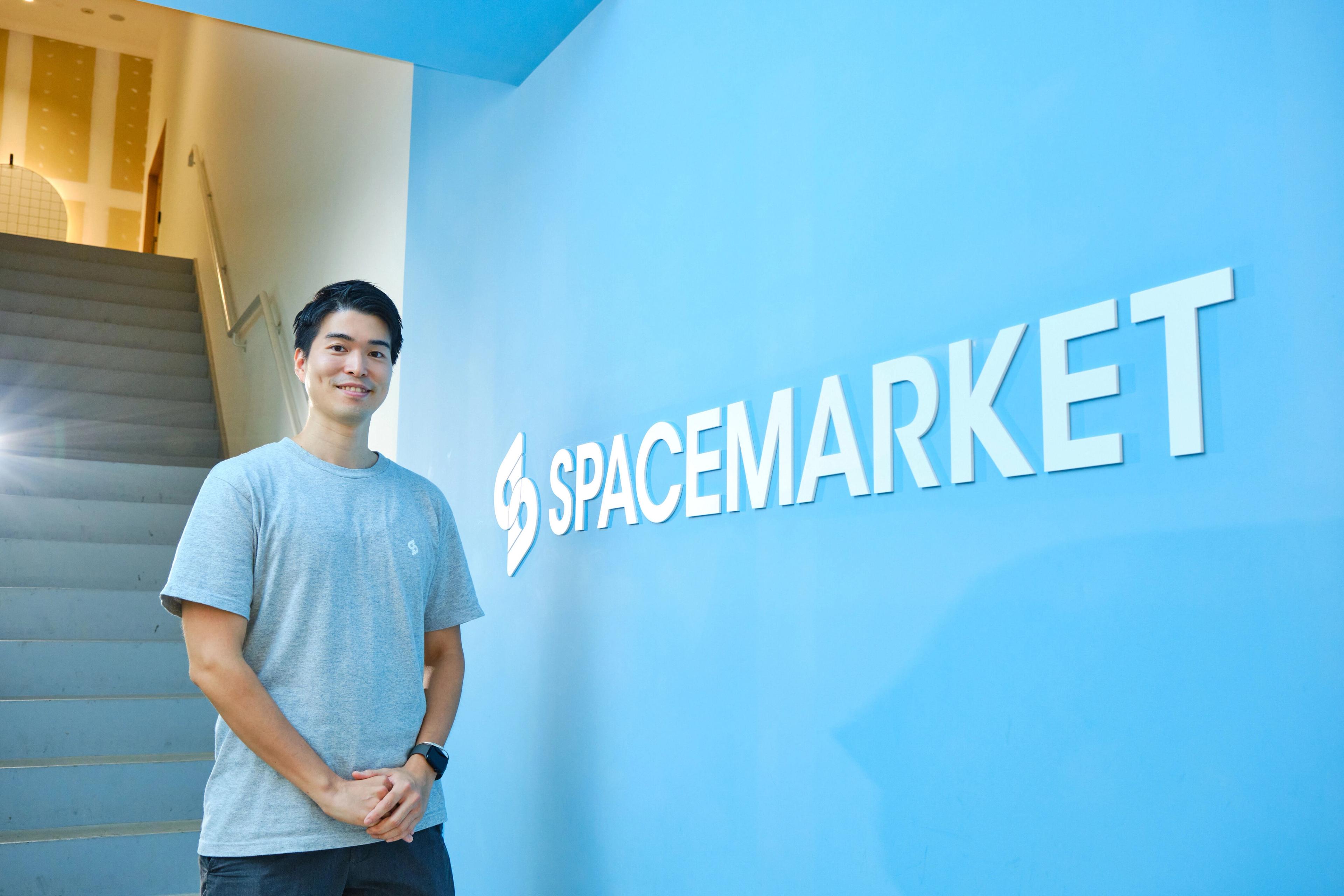 青い壁の前に立つ男性が、"SPACEMARKET"のロゴの横にいます。階段が背景に見えます。