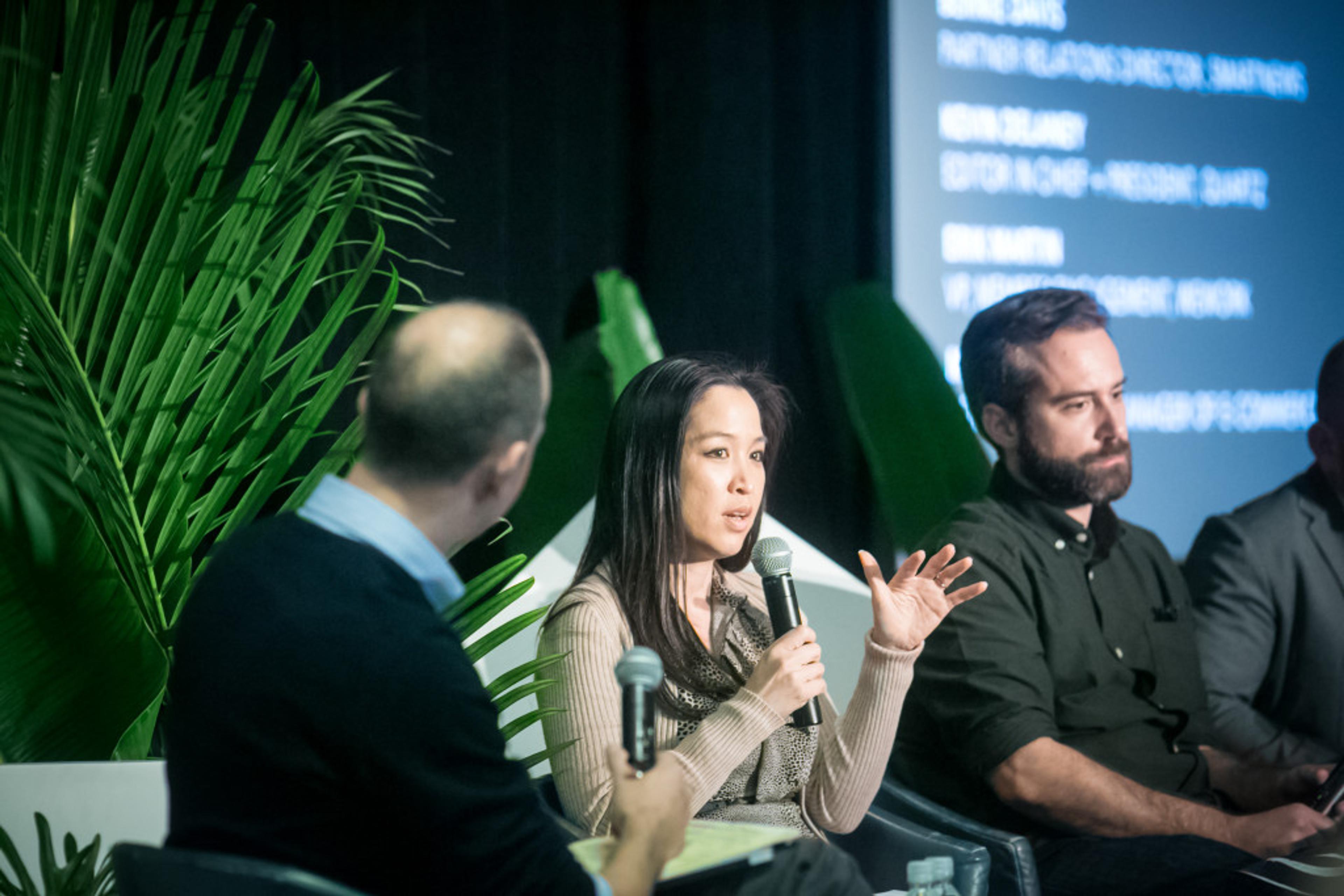 A woman speaks into a microphone during a panel discussion, while two men listen attentively. The background features large green leaves and a digital screen displaying information.