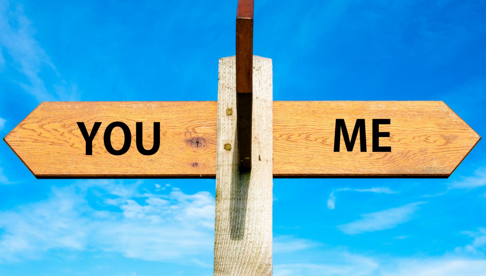 A wooden signpost with two arrows pointing in opposite directions, labeled "YOU" on one side and "ME" on the other, against a blue sky.