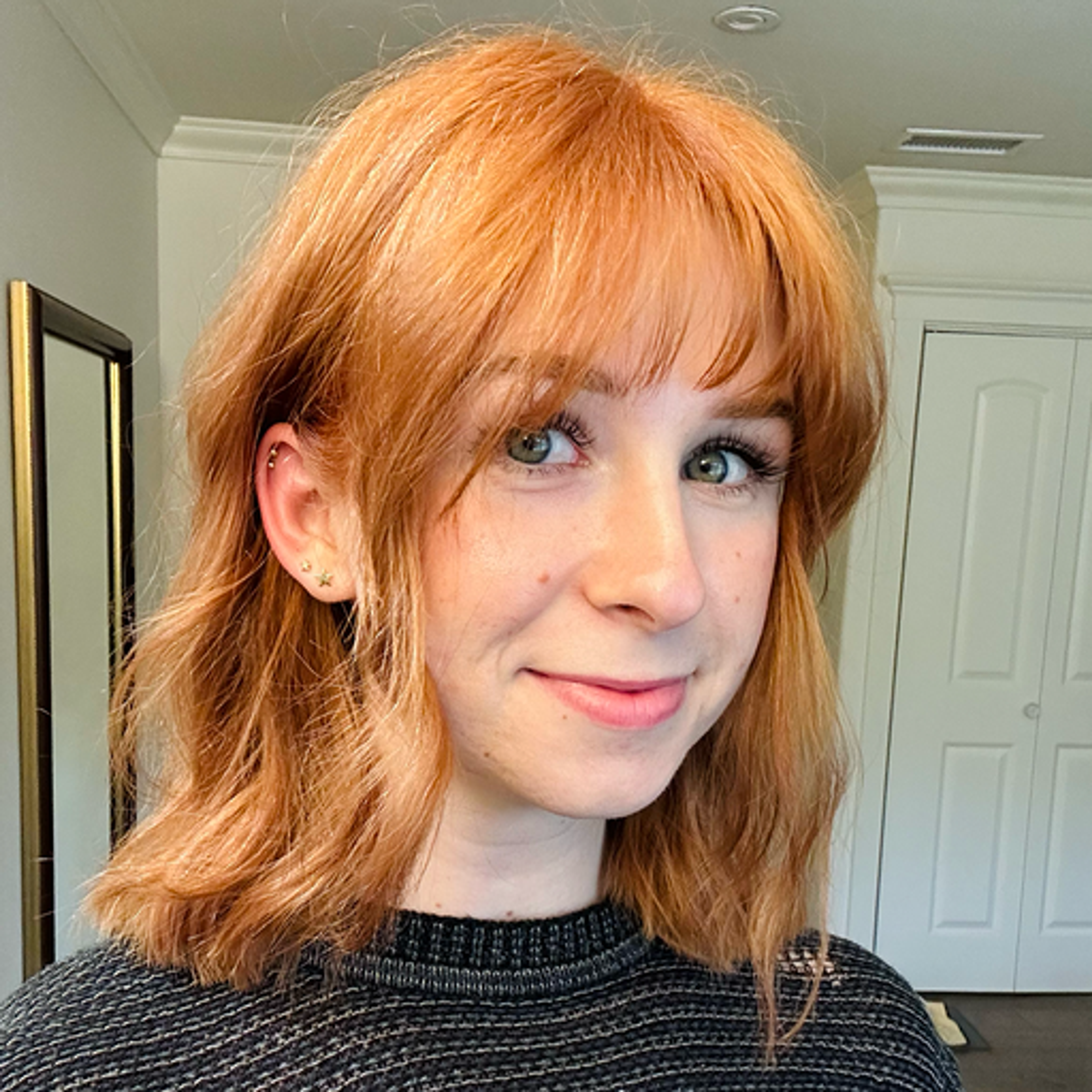 A young woman with wavy, shoulder-length reddish hair smiles at the camera. She is wearing a dark sweater and standing in a well-lit room with a mirror in the background.
