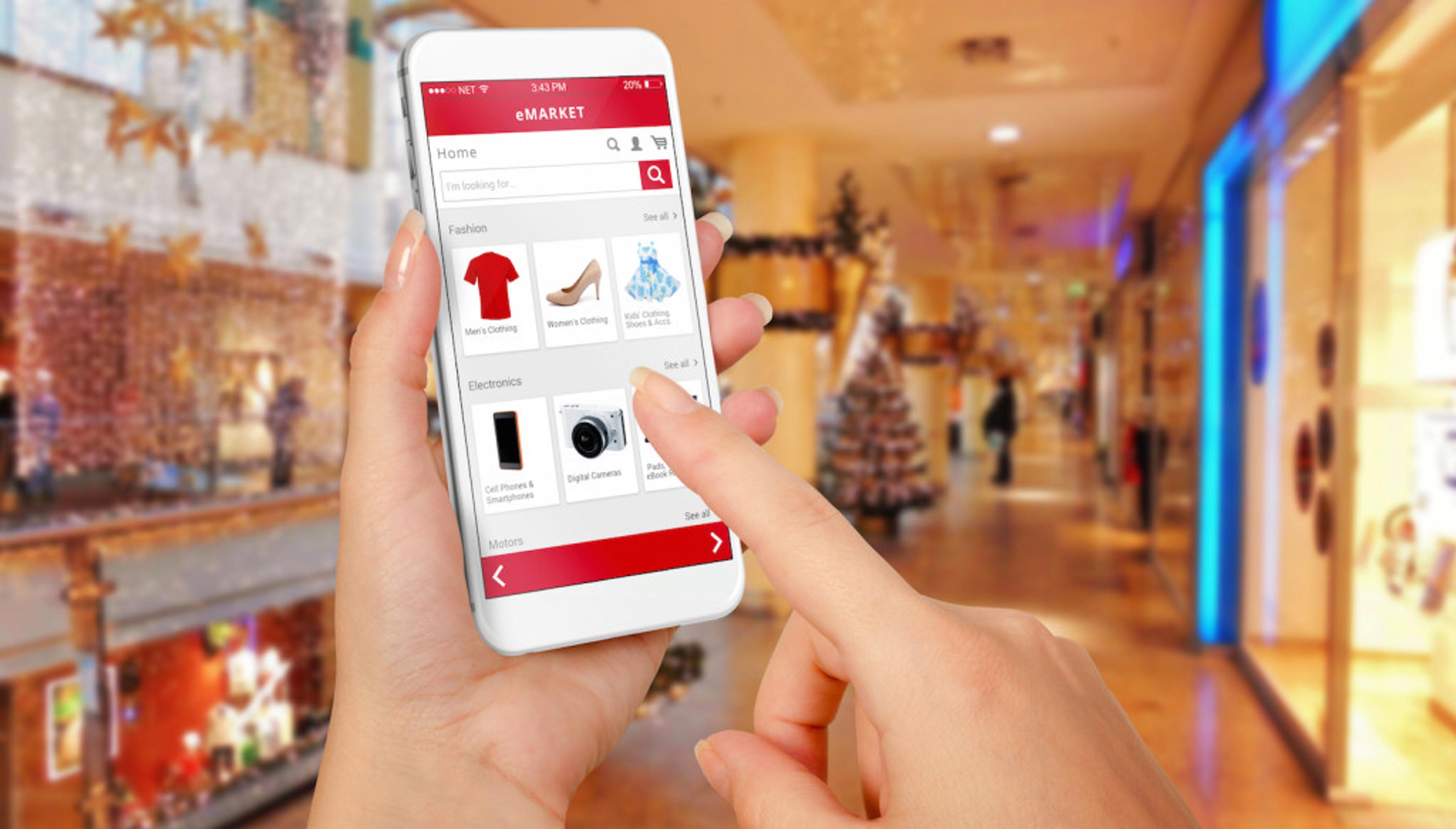 A person using a smartphone to browse an online shopping app in a festive shopping mall. The screen displays various product categories like fashion and electronics.