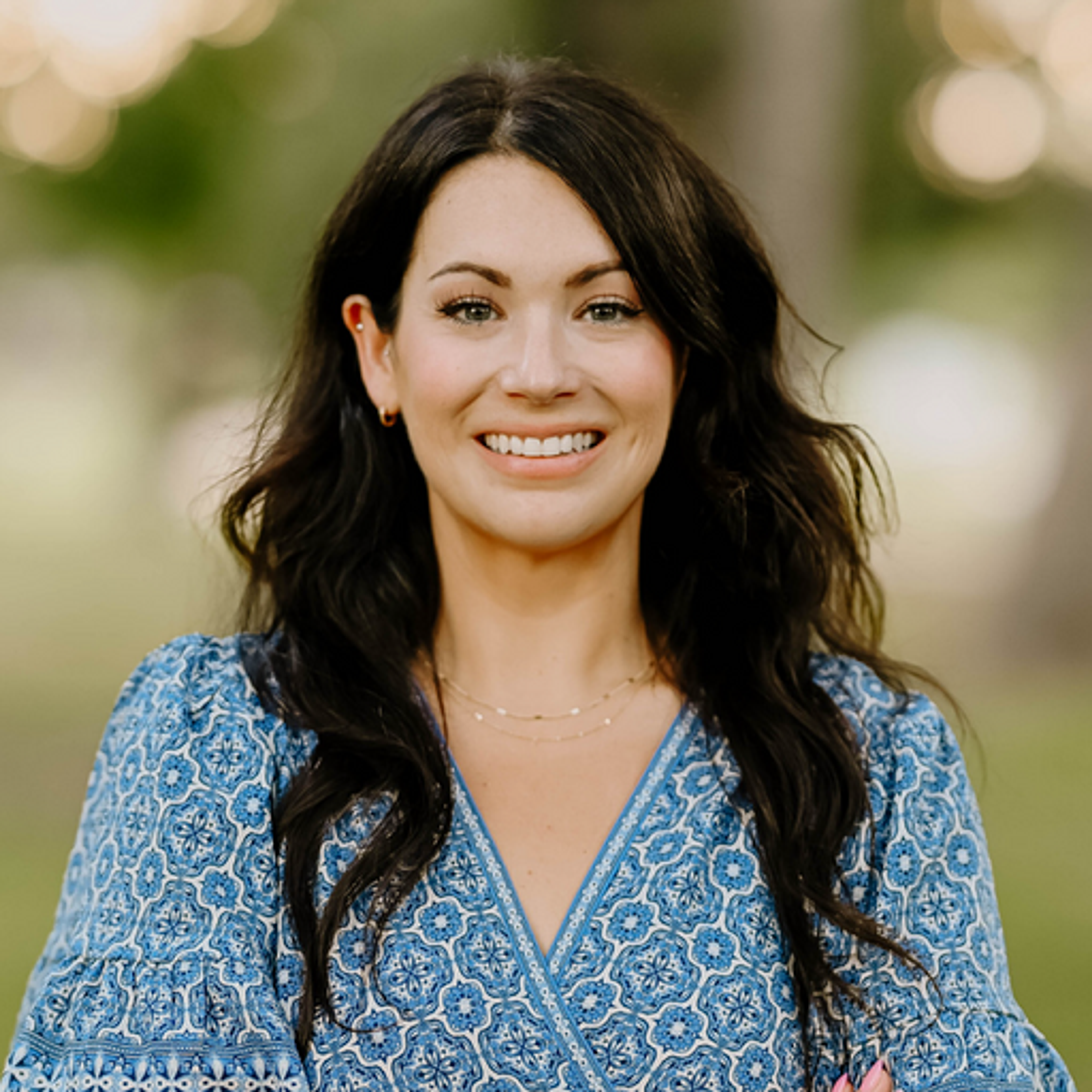 Smiling woman with long dark hair wearing a navy top, posing in front of a softly blurred light background.