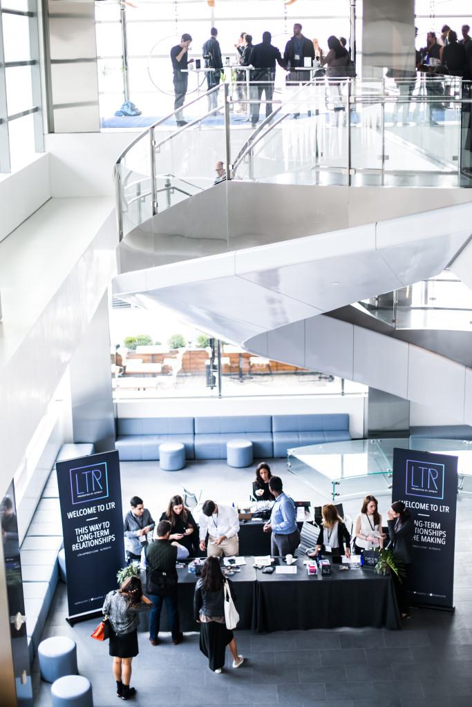 A busy conference registration area with attendees mingling and a welcome booth displaying "LTR" banners. The space features modern architecture and a staircase in the background.