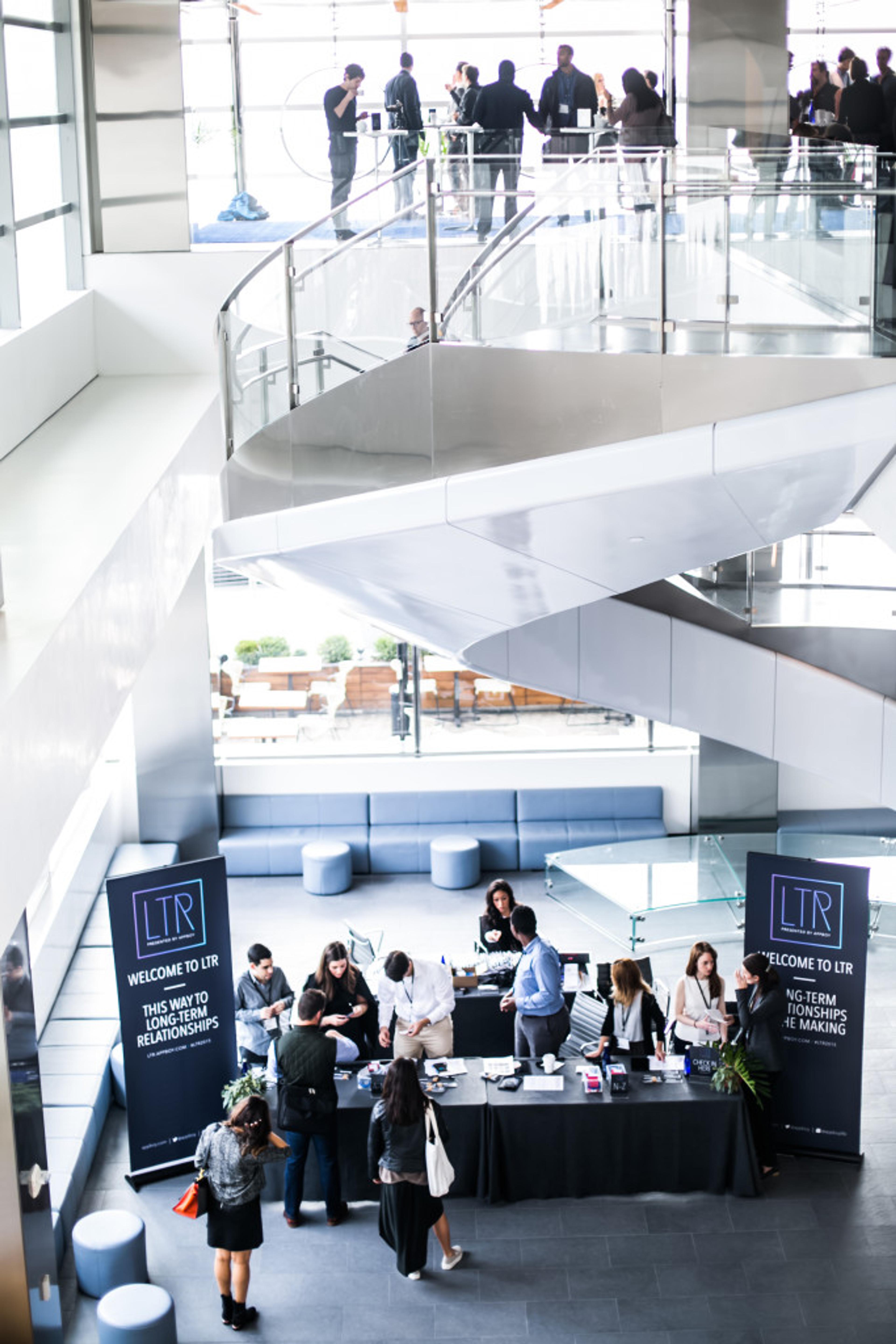 A busy conference registration area with attendees mingling and a welcome booth displaying "LTR" banners. The space features modern architecture and a staircase in the background.