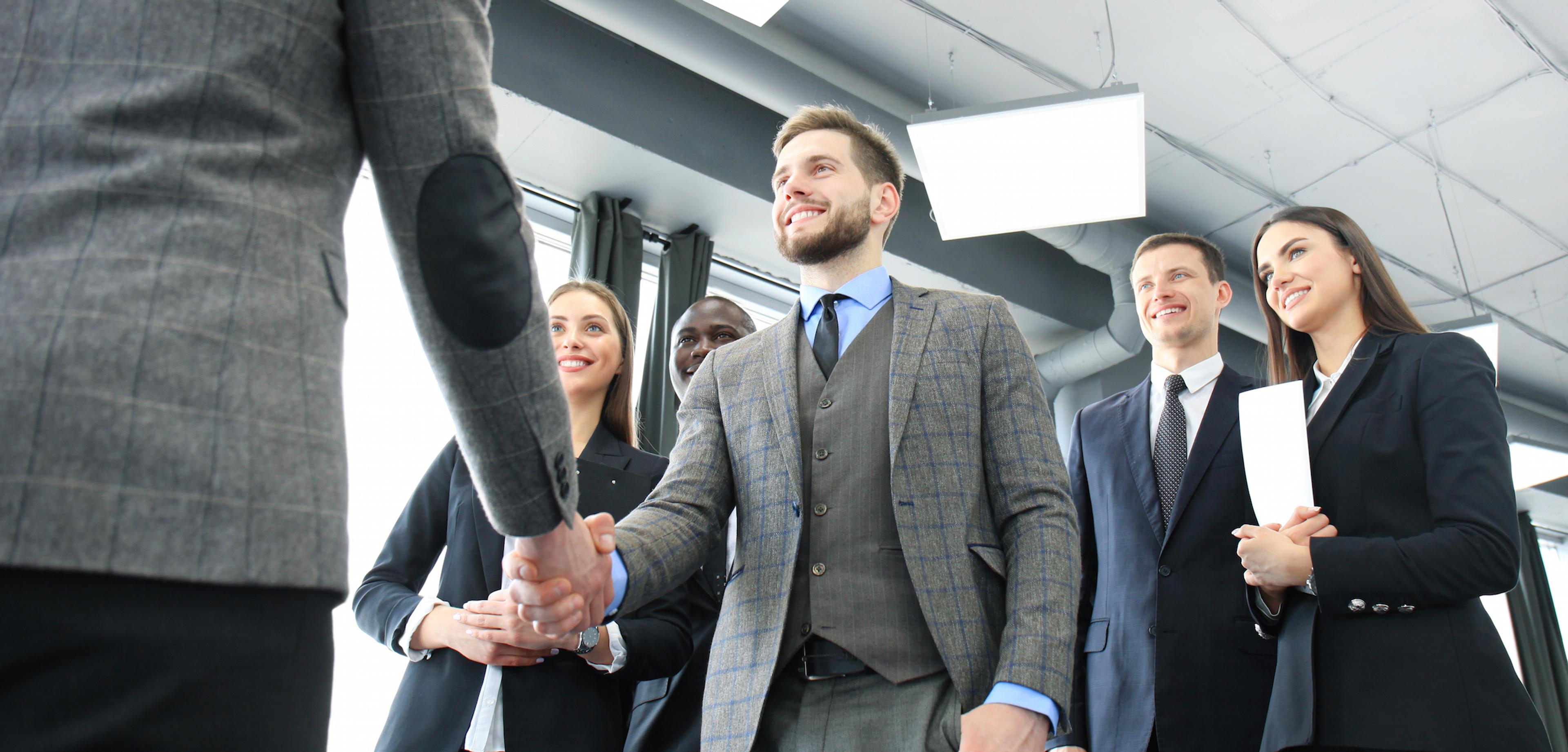 A group of professionals in business attire are smiling and shaking hands in an office setting. The atmosphere is positive and collaborative.