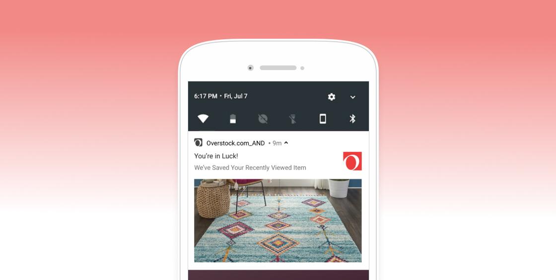 A smartphone screen displaying a notification from Overstock.com about a recently viewed item, featuring a colorful area rug in a room setting. The background is a gradient from pink to white.