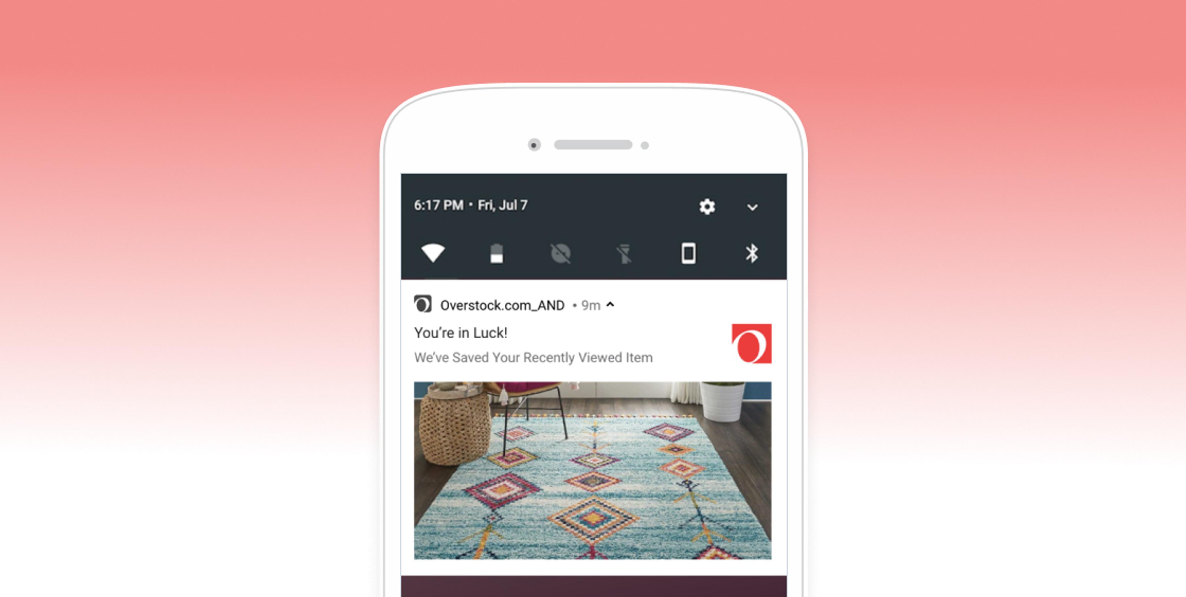 A smartphone screen displaying a notification from Overstock.com about a recently viewed item, featuring a colorful area rug in a room setting. The background is a gradient from pink to white.