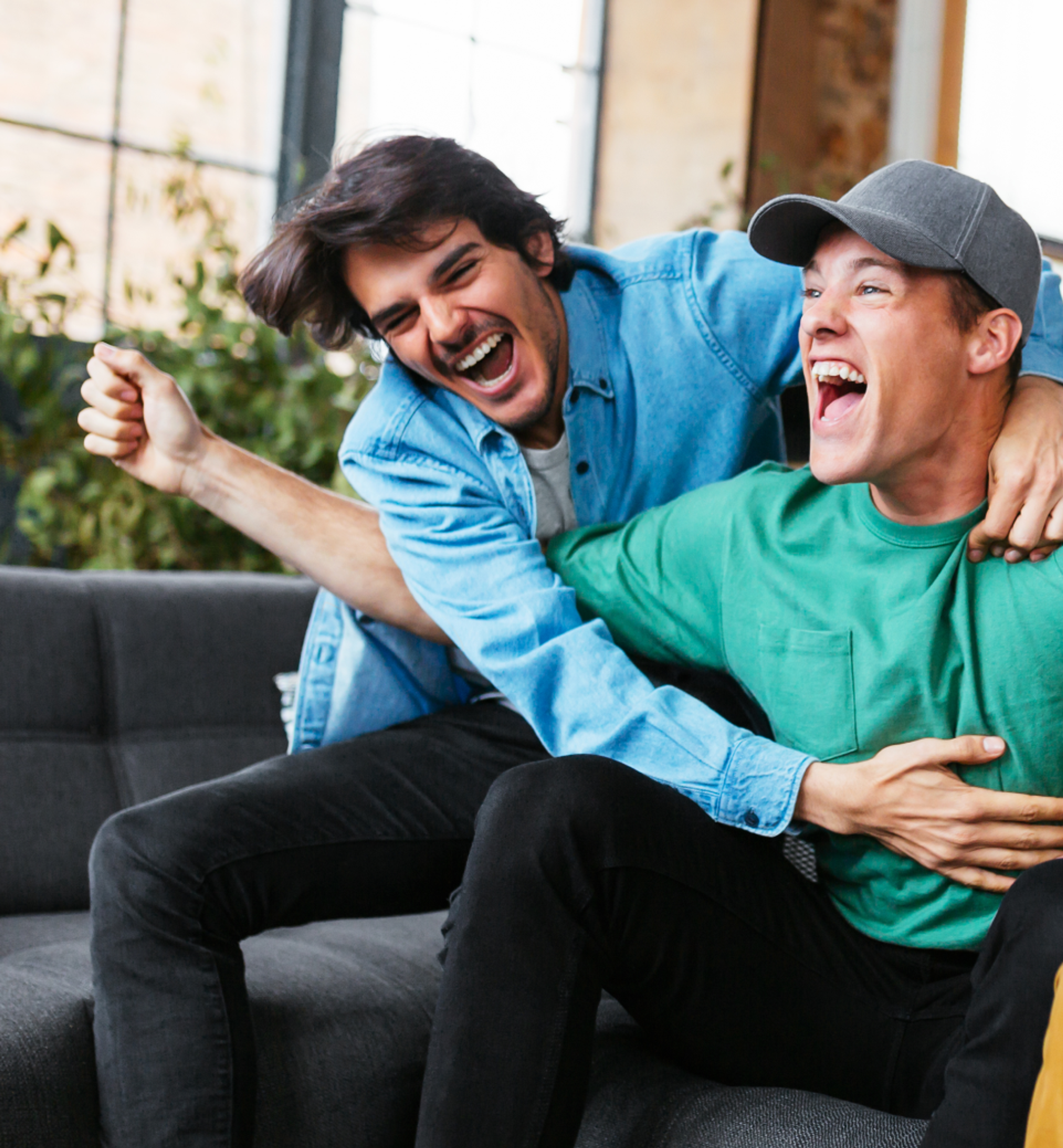 Two friends sitting on a couch laugh and hug each other enthusiastically.