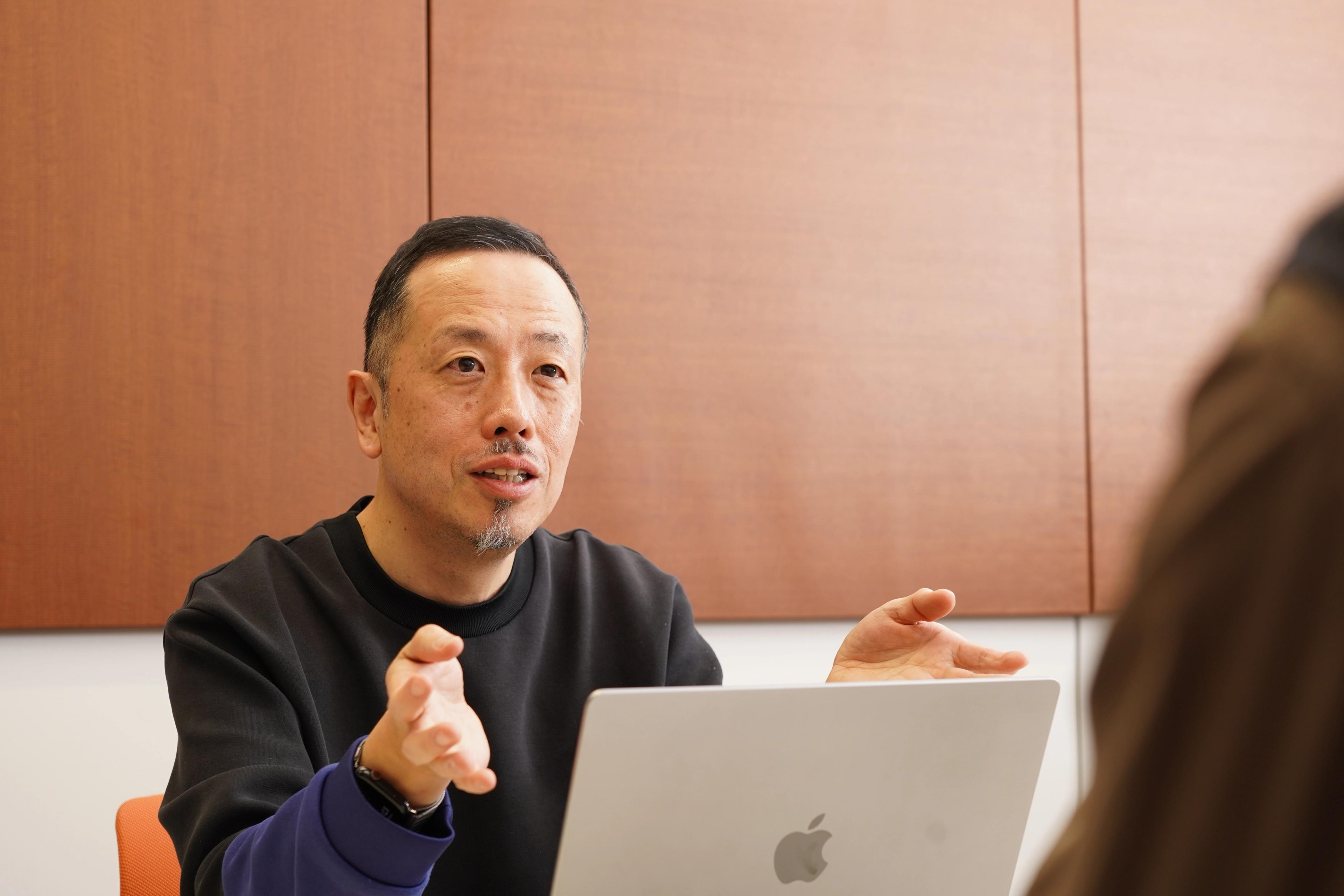 あごひげを生やした東アジア系の男性が話しながら身振り手振りをし、目の前のテーブルにはノートパソコンが置かれている。