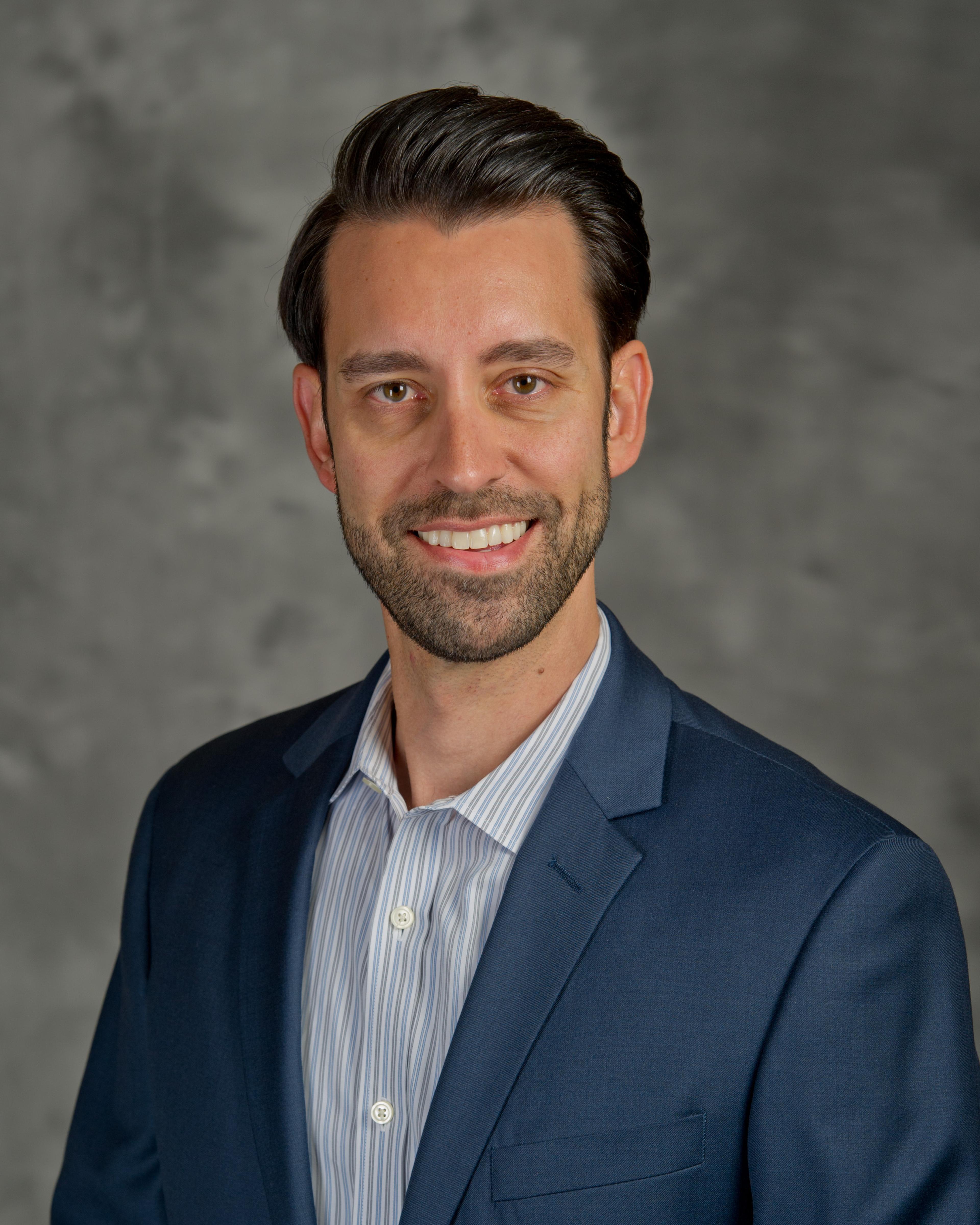 A man in a blue suit jacket and striped shirt smiles at the camera against a gray background. He has dark hair styled neatly.