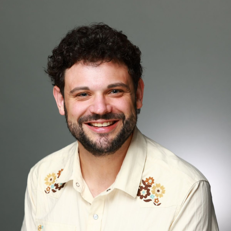 Headshot of a smiling man with curly dark hair and a beard, wearing a cream shirt with floral embroidery.