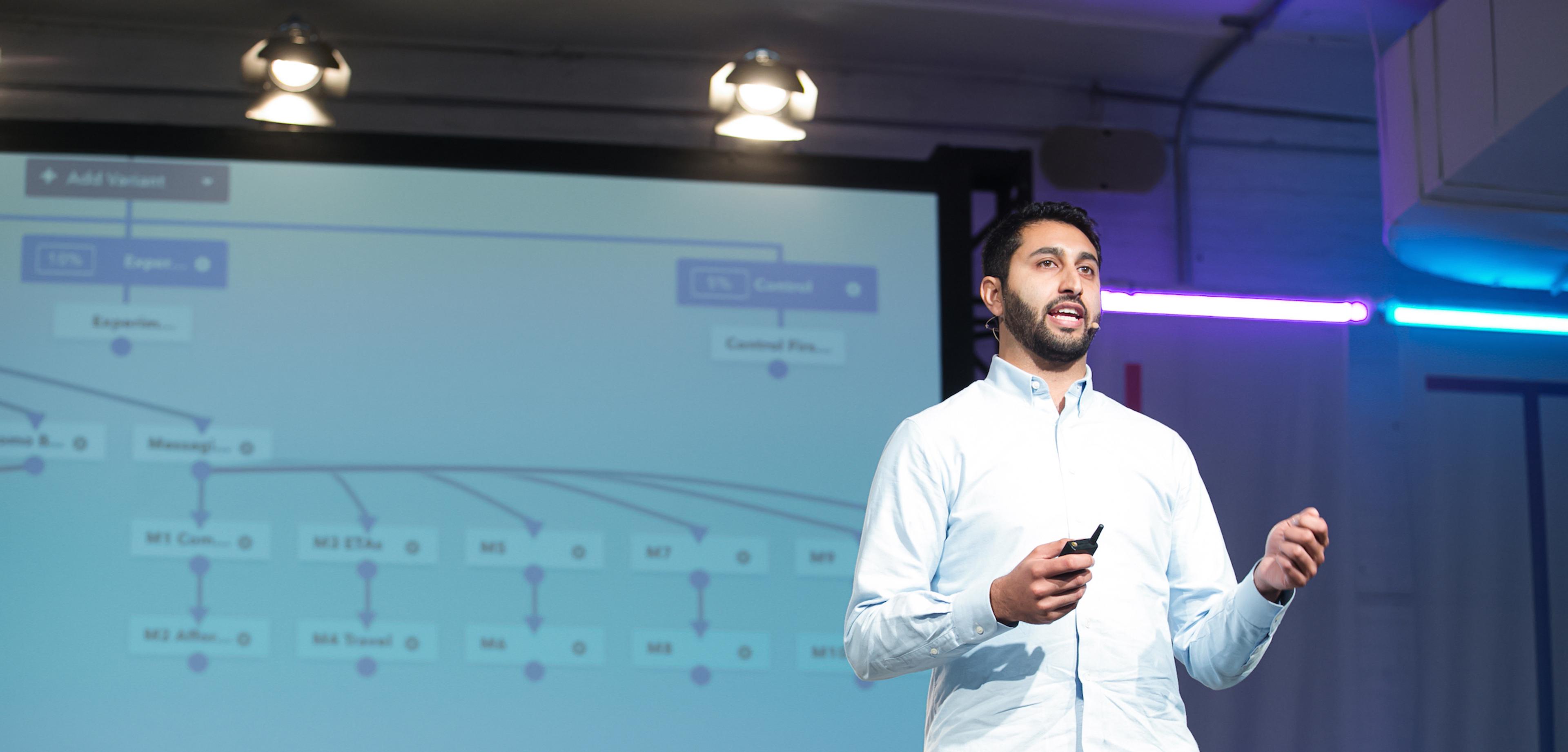 A man is speaking on stage with a presentation slide behind him displaying a flowchart. He is holding a microphone and gesturing as he presents.