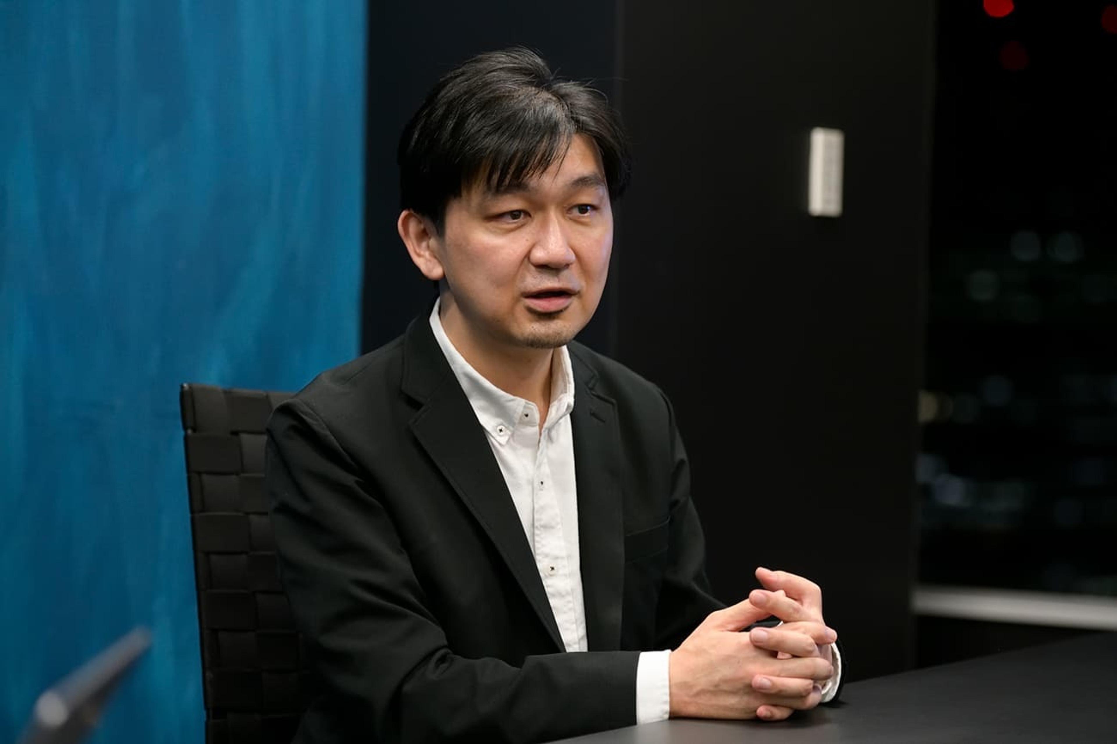 黒いスーツを着た男性がテーブルに座っている。背景には青い壁がある。