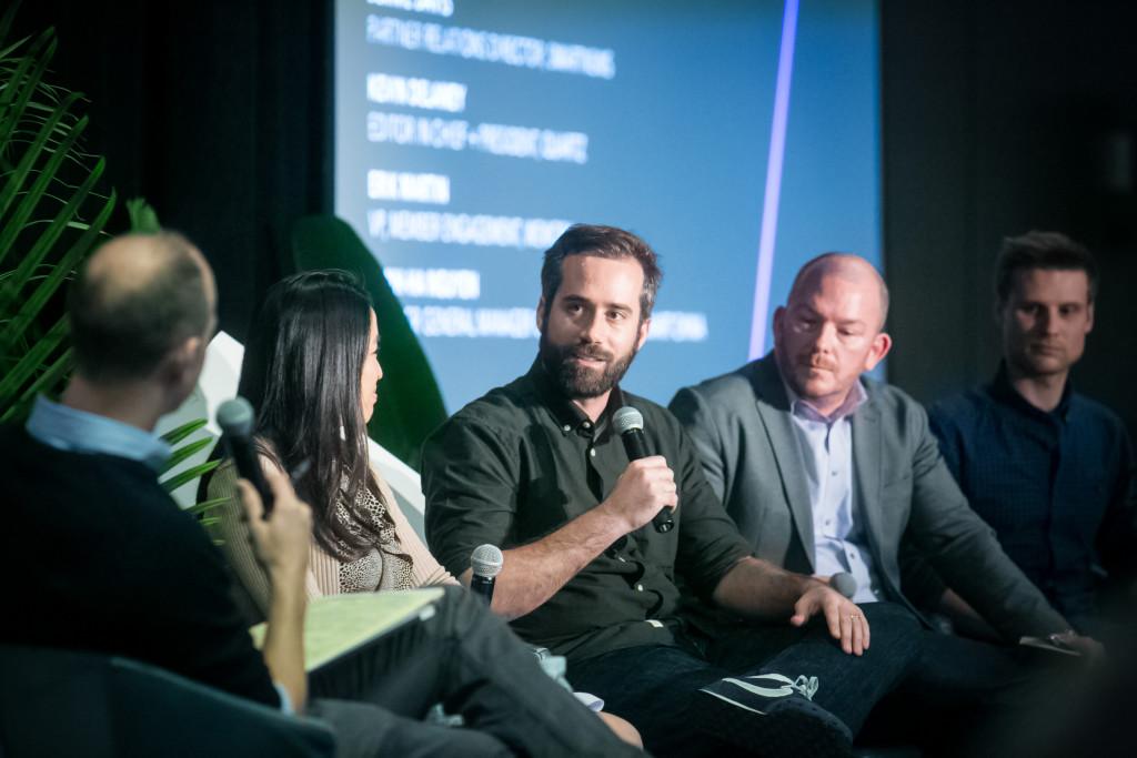 A panel discussion featuring four speakers, with one man speaking into a microphone. The setting includes a dark background and plants in the foreground.