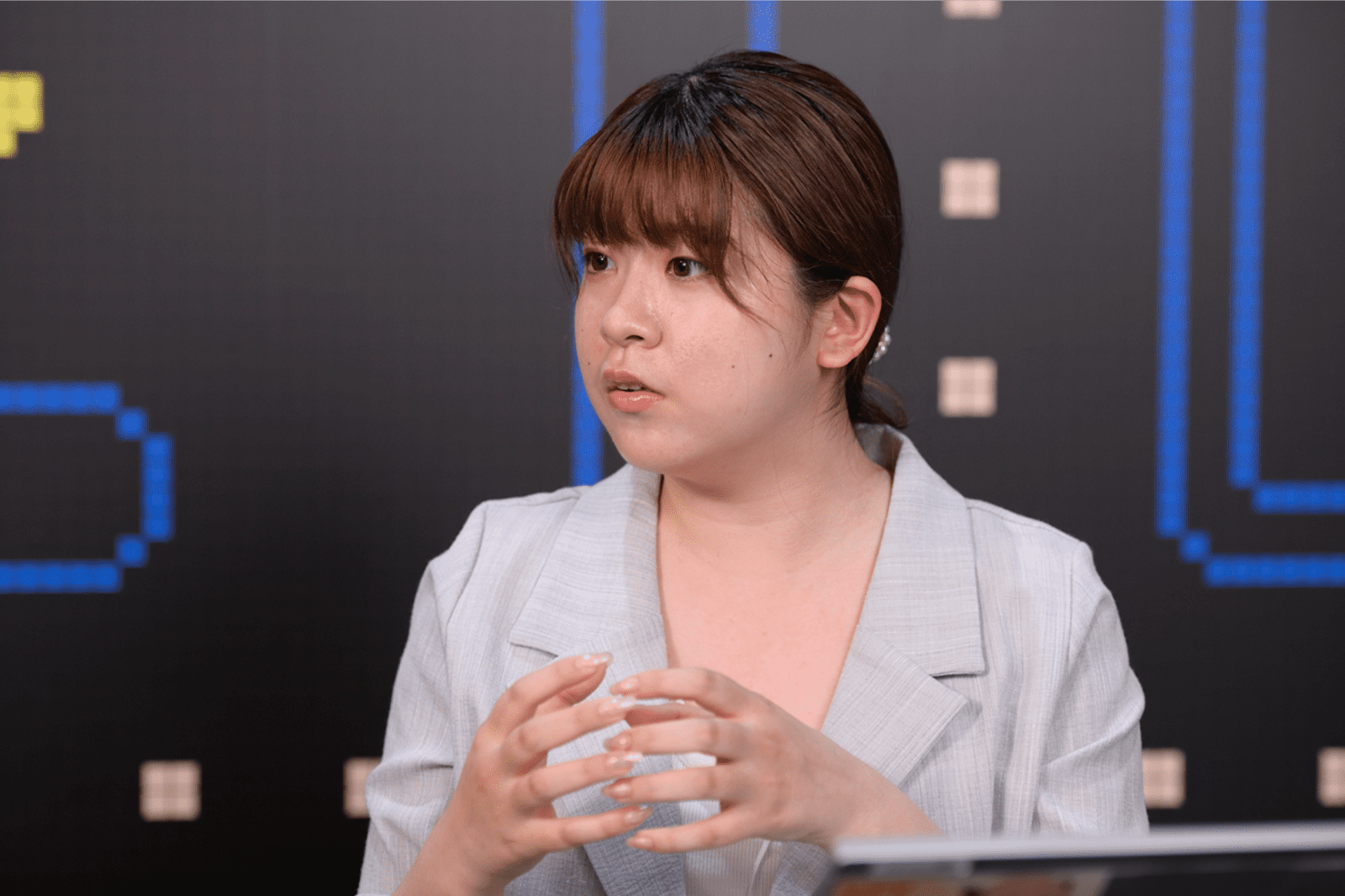 バンダイナムコ社員の一人が、ゲーム画面の背景の前で腕をくんで座ってインタビューに答えている様子