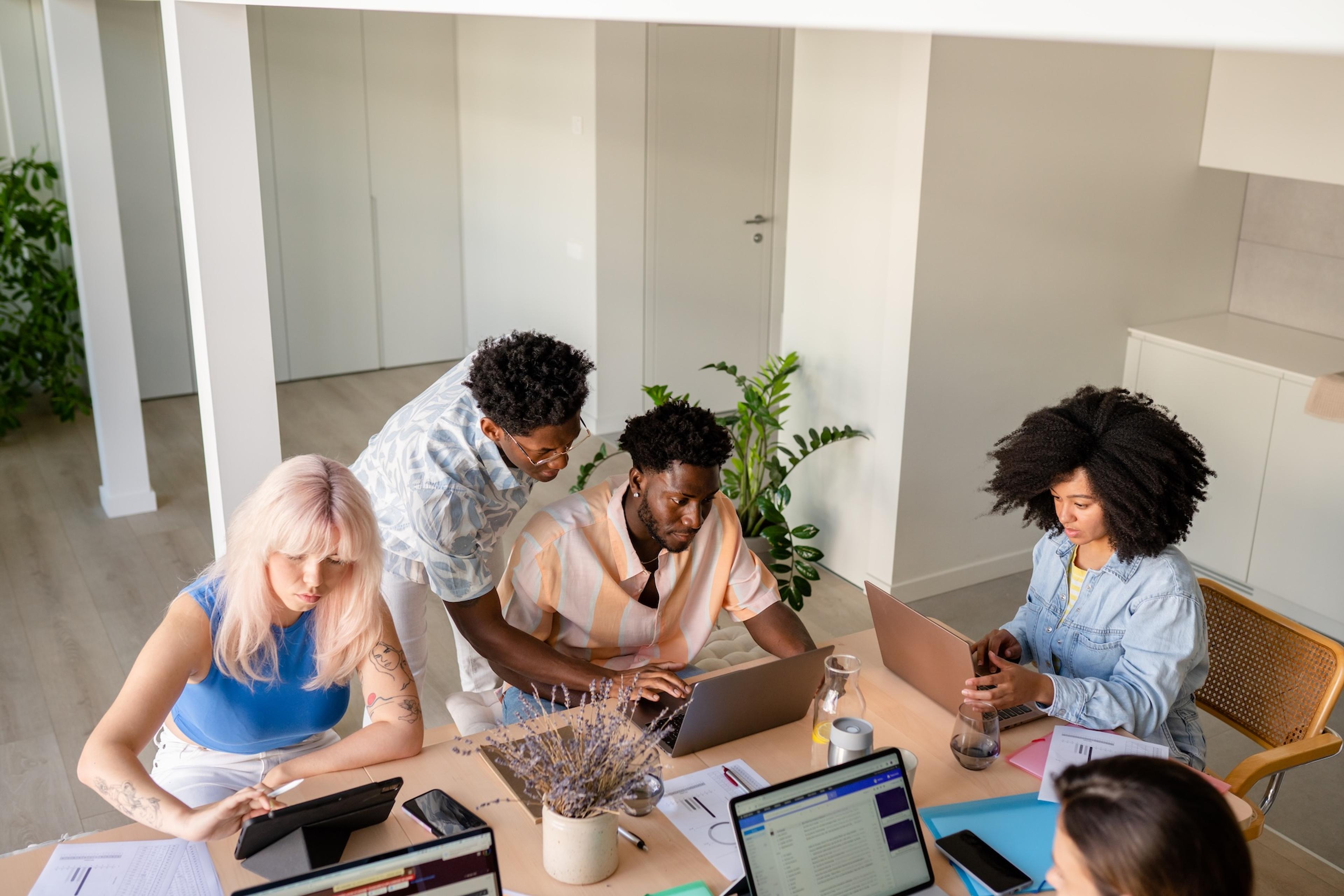 A diverse group of people working on laptops and tablets around a table in a modern office.