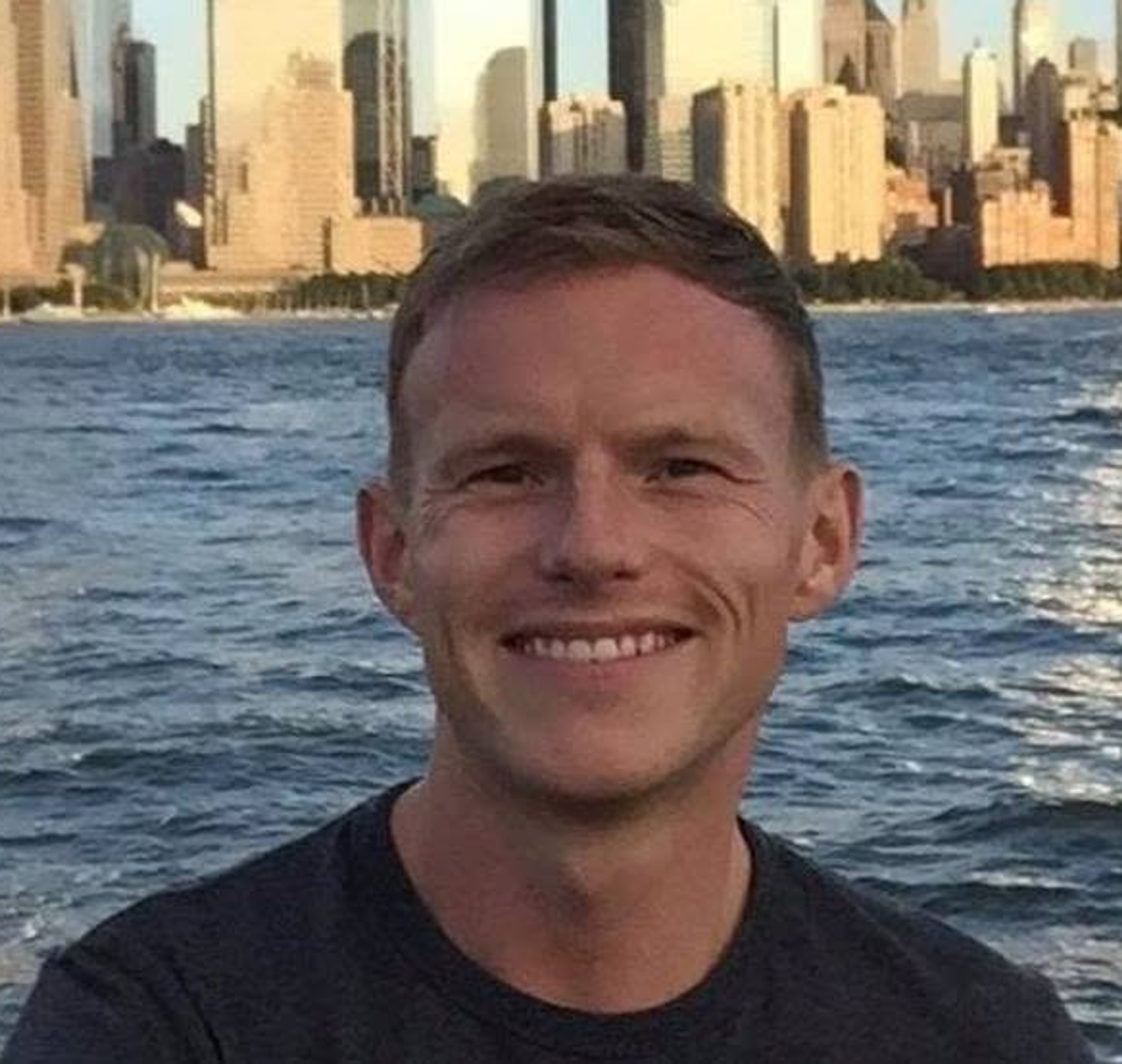 Smiling person in a dark T-shirt standing by the water with a city skyline in the background.