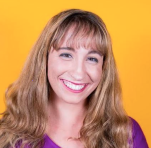 A smiling woman with long, wavy brown hair stands against a bright yellow background. She is wearing a purple top and has a cheerful expression.