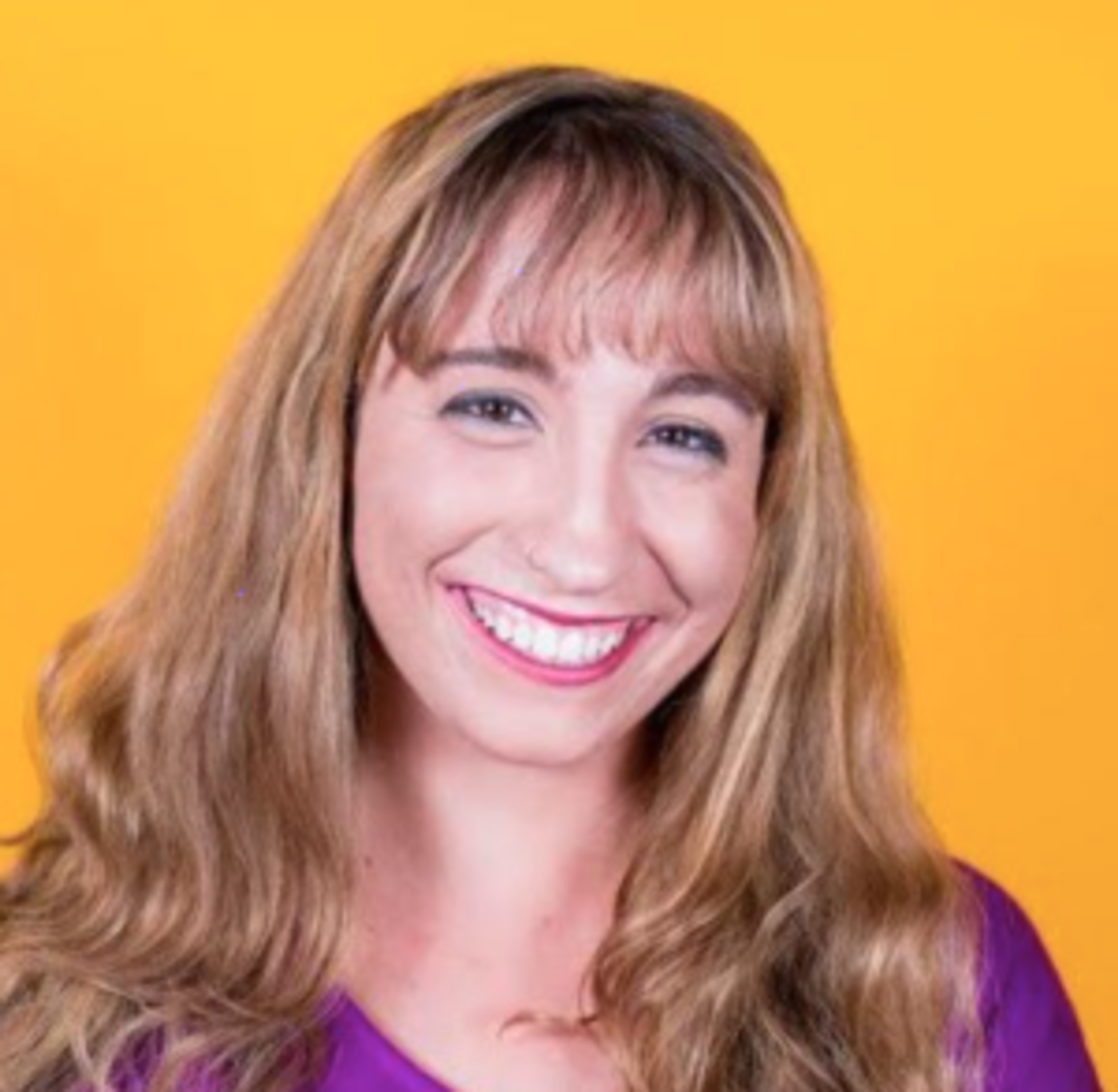 A smiling woman with long, wavy brown hair stands against a bright yellow background. She is wearing a purple top and has a cheerful expression.