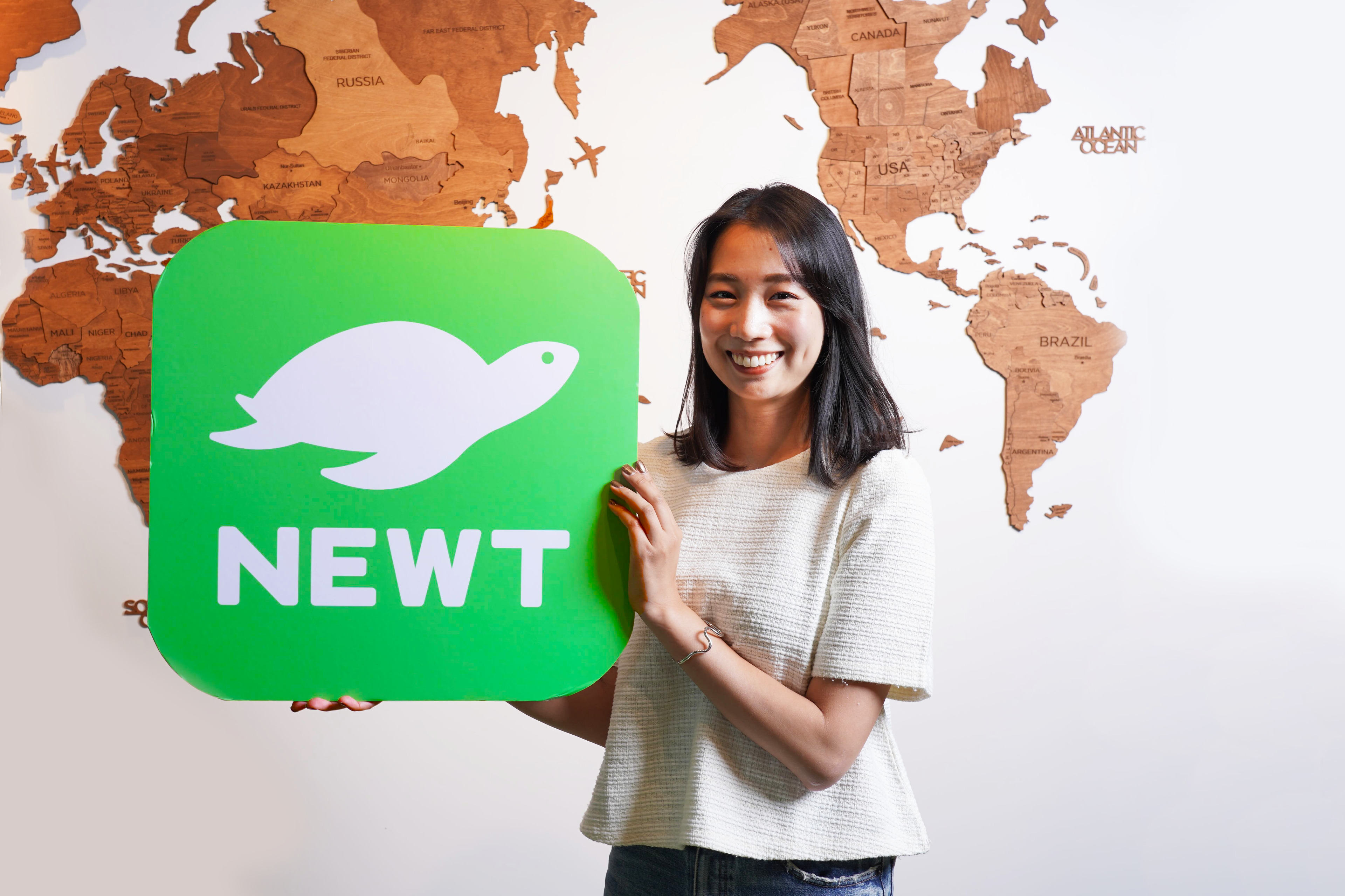 女性が「NEWT」というロゴを持って微笑んでいる。背景には世界地図が描かれている。
