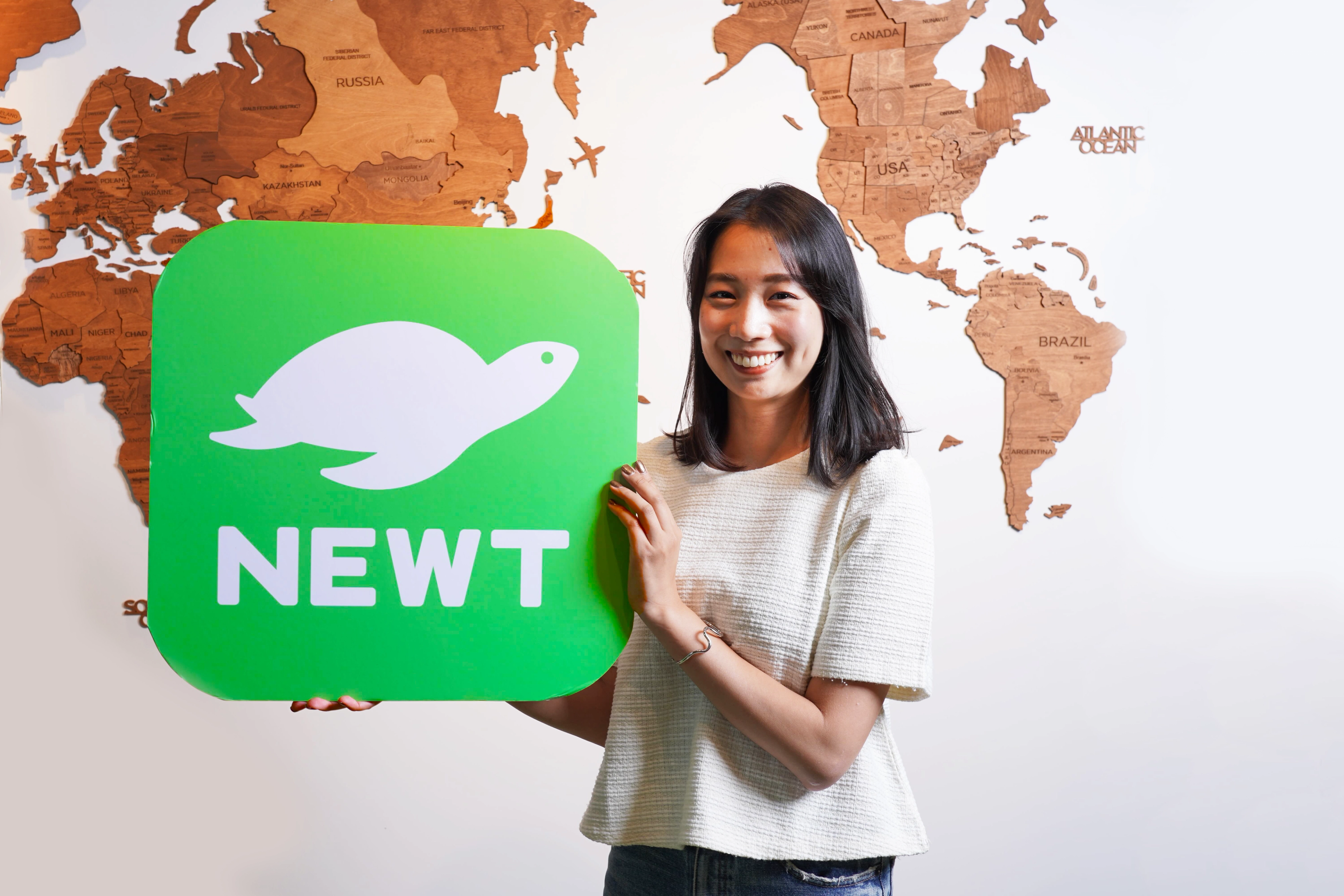 女性が「NEWT」というロゴを持って微笑んでいる。背景には世界地図が描かれている。