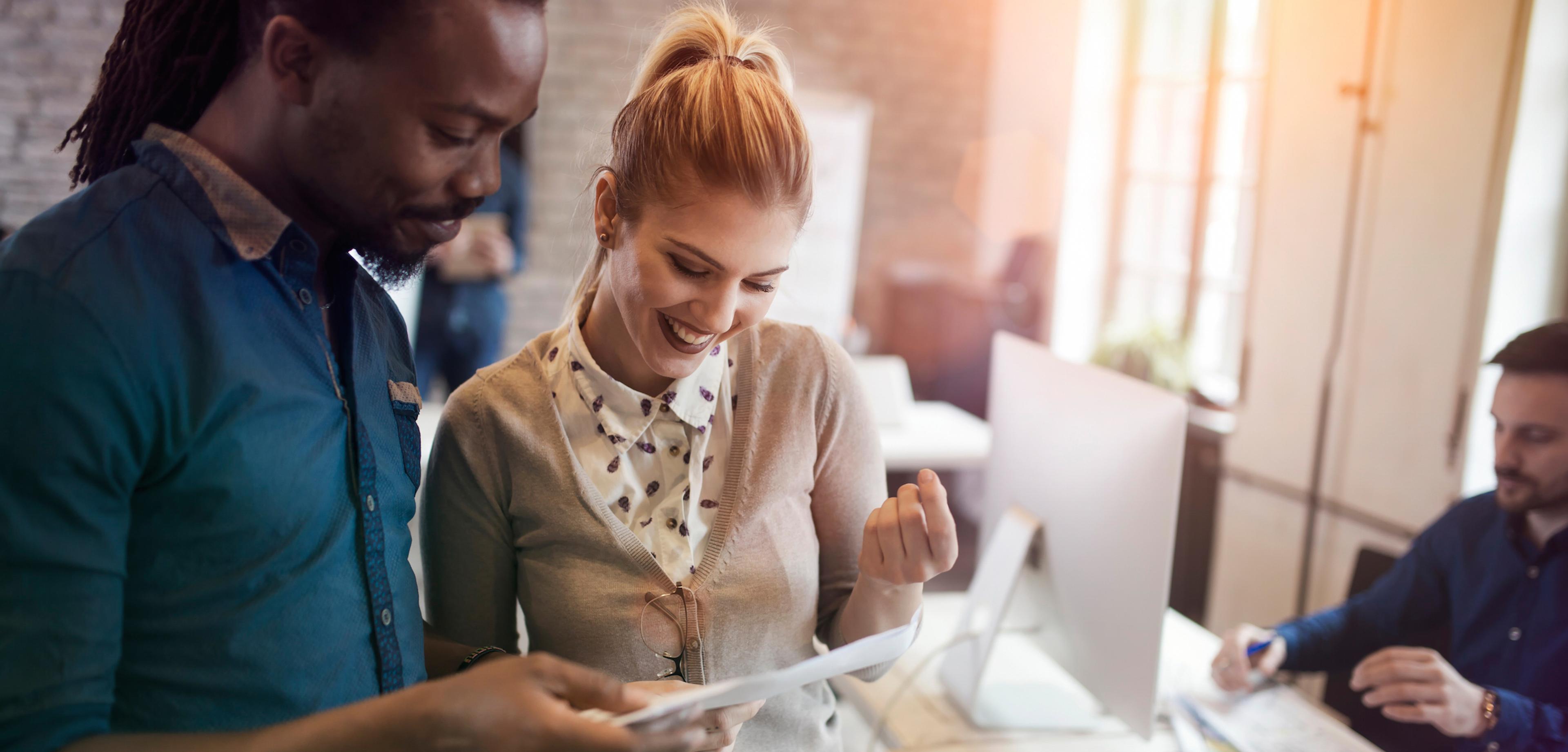 A man and a woman are looking at a document together, smiling and engaged in conversation. In the background, another person is working at a desk.