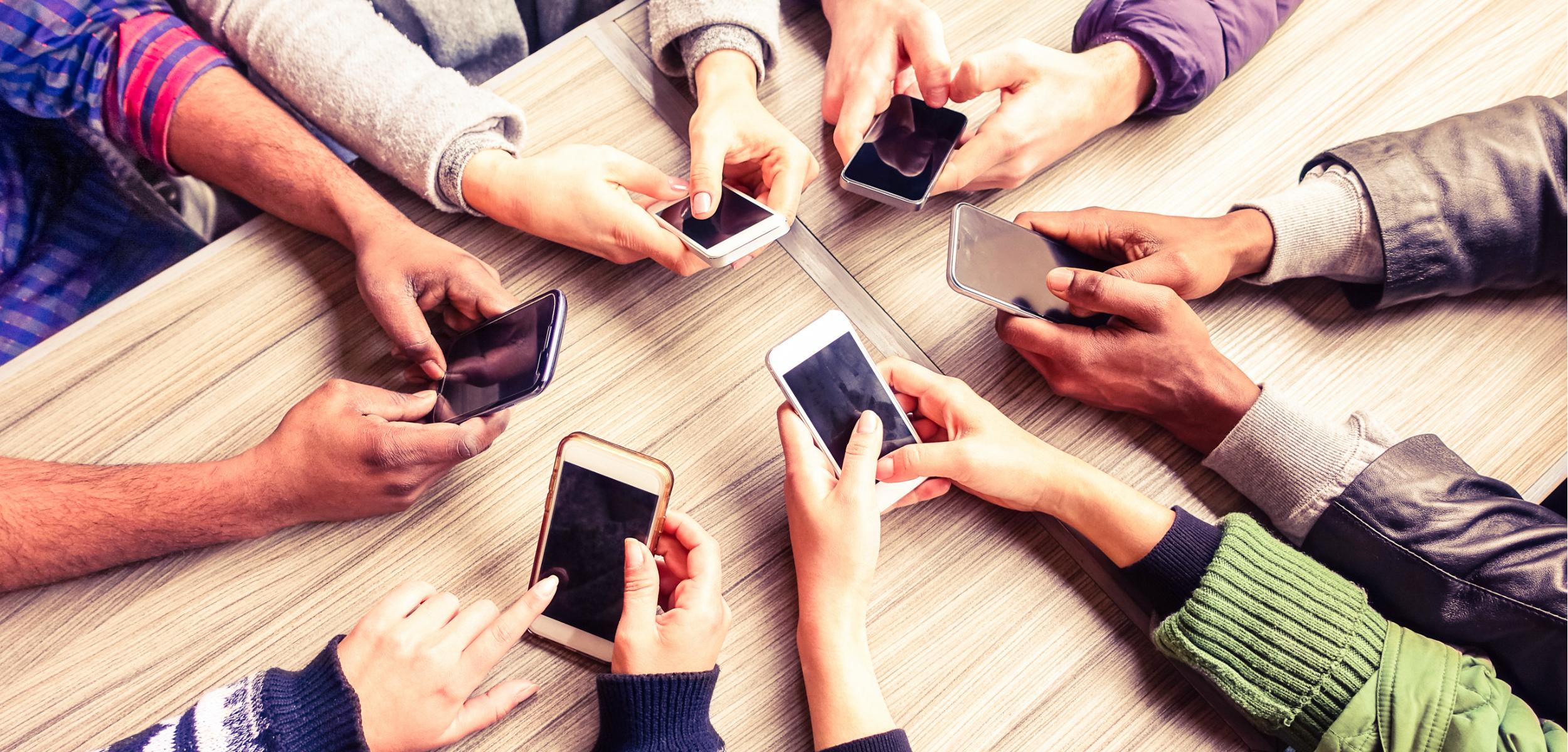 A group of people holding smartphones, gathered around a table. Their hands are visible, with various phone models displayed.