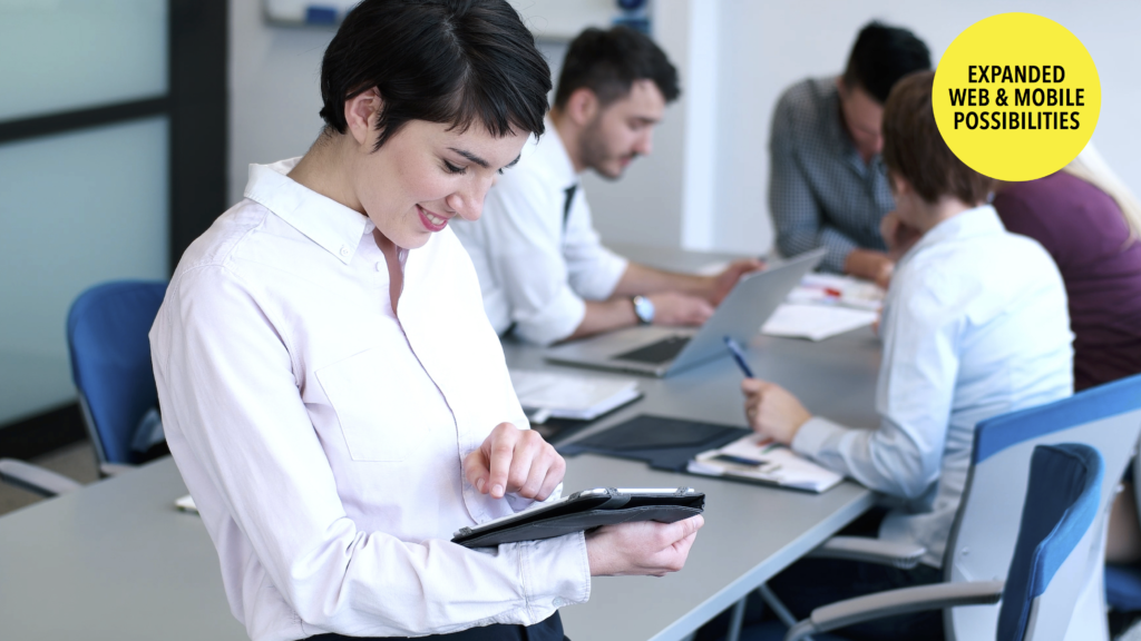 A woman in a white shirt smiles while using a tablet in a meeting room. In the background, several people are engaged in discussion around a table with laptops and documents.