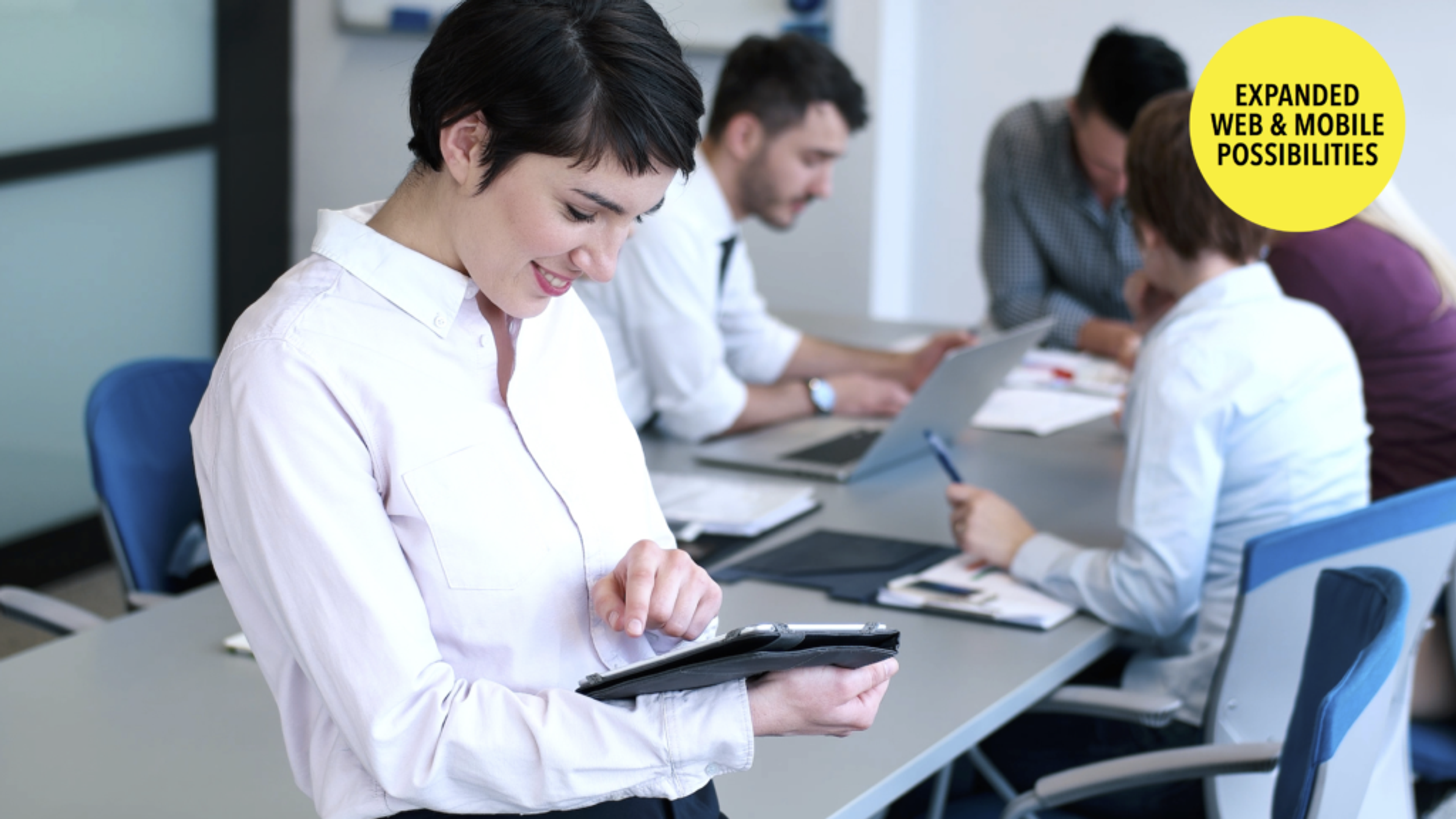 A woman in a white shirt smiles while using a tablet in a meeting room. In the background, several people are engaged in discussion around a table with laptops and documents.