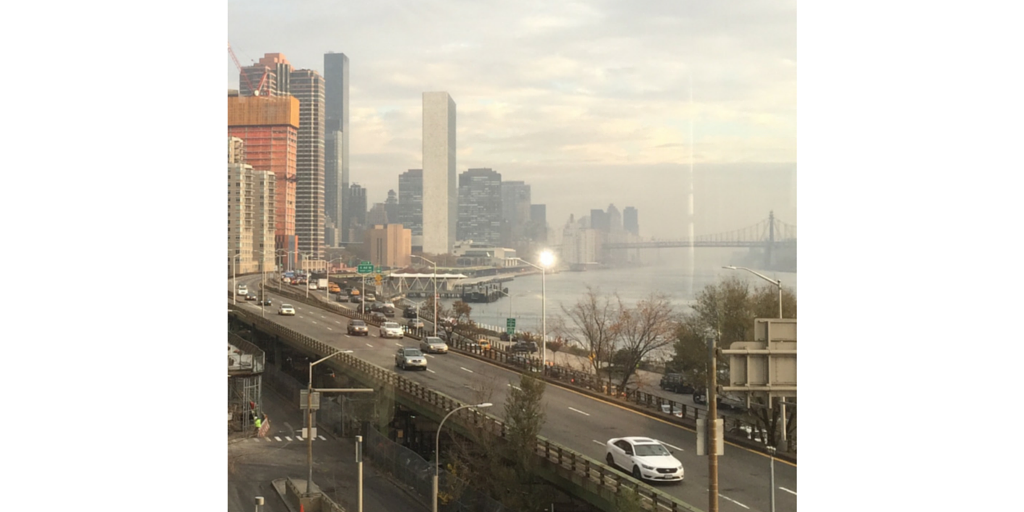 A view of a city skyline with tall buildings along a waterfront, featuring a highway with cars and a bridge in the distance. The scene is set under a cloudy sky.