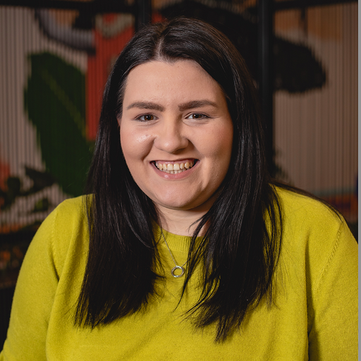 A smiling woman with long dark hair wearing a bright yellow sweater. She is in an indoor setting with plants in the background.