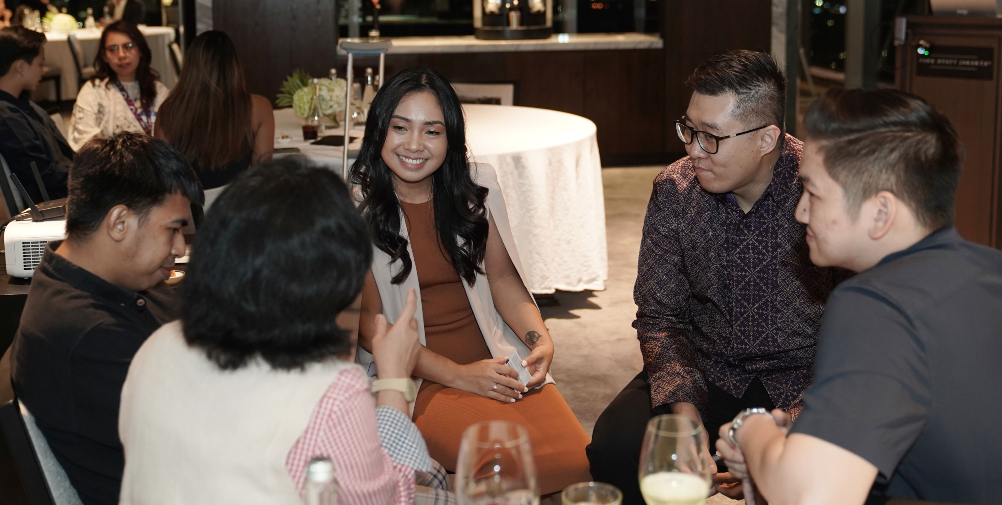 A group of five people engaged in conversation at a social gathering, seated around a table in a well-lit setting. The atmosphere appears relaxed and friendly.