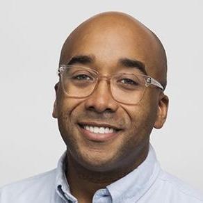 A smiling man with a bald head and glasses, wearing a light blue shirt. He has a friendly expression against a plain background.