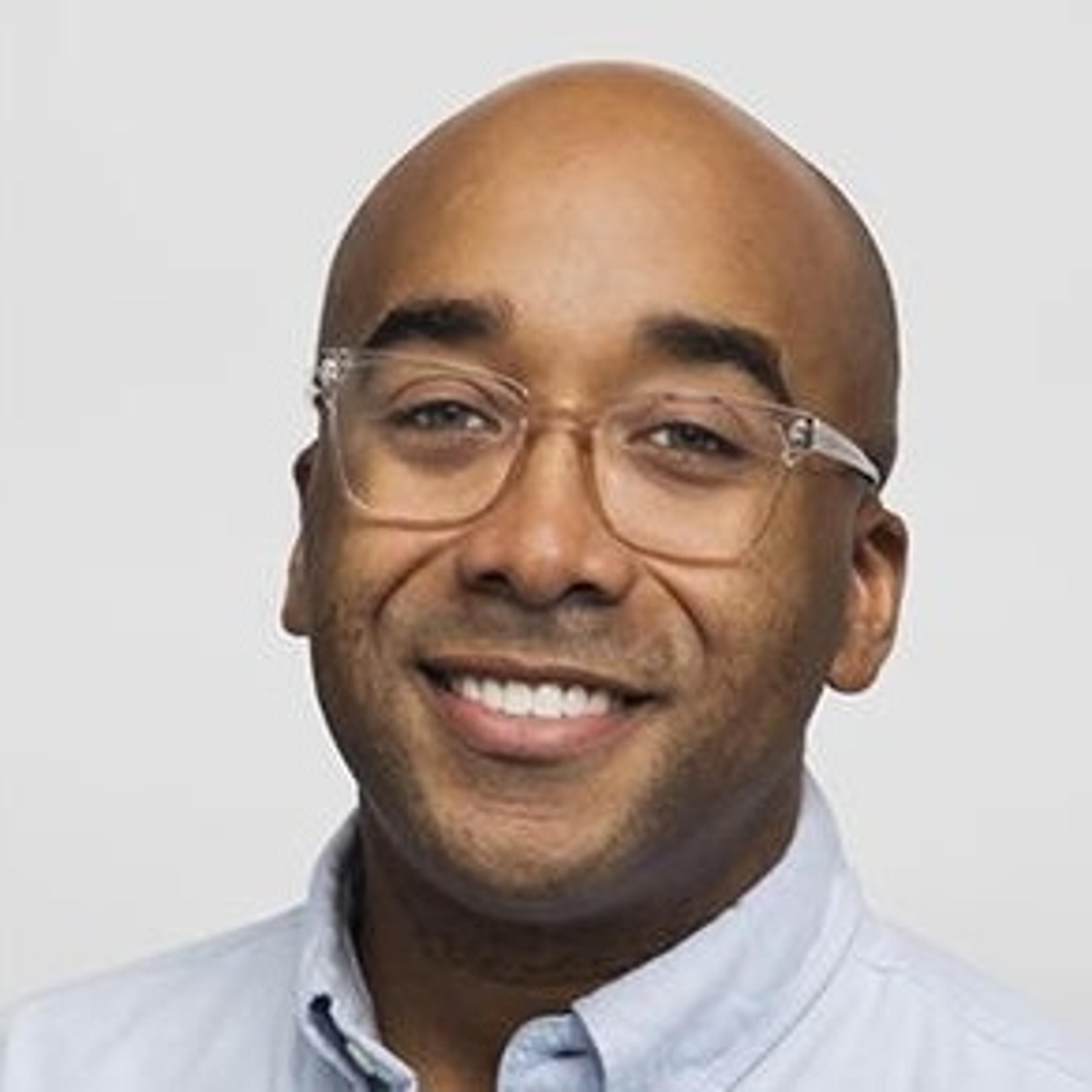 A smiling man with a bald head and glasses, wearing a light blue shirt. He has a friendly expression against a plain background.