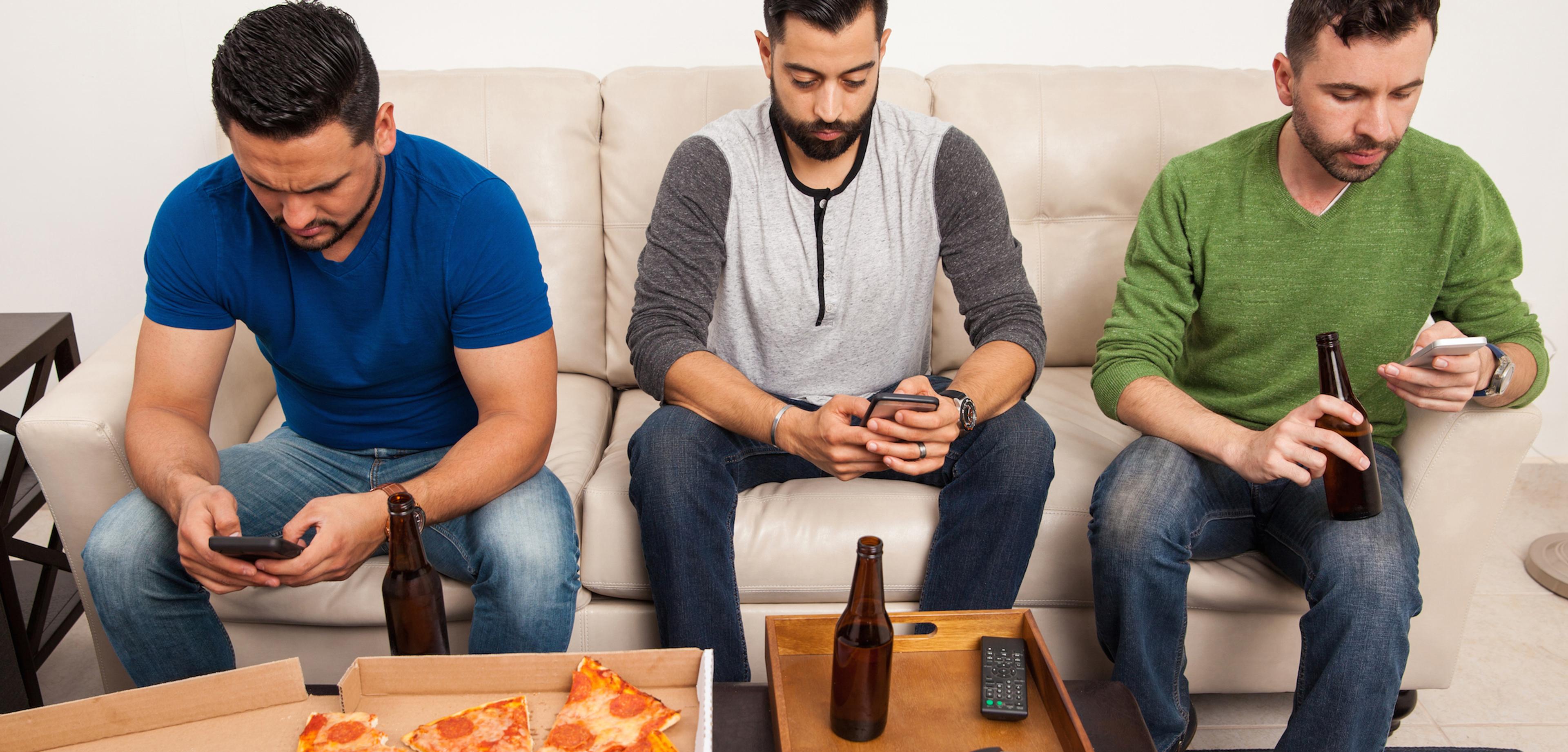 Three men sitting on a couch, each focused on their smartphones. A pizza box and drinks are on the table in front of them.