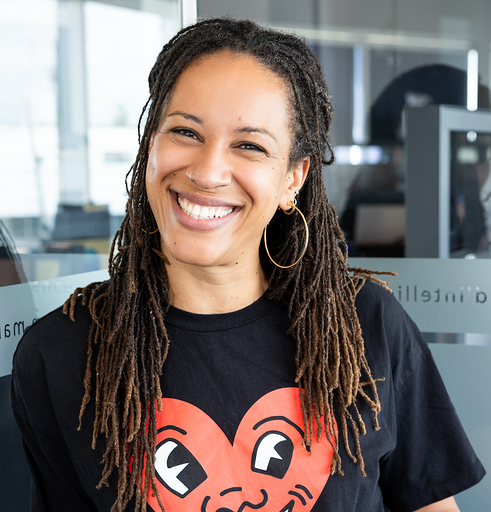 Headshot of a smiling woman with long locs and hoop earrings, wearing a black T-shirt with a red heart graphic.