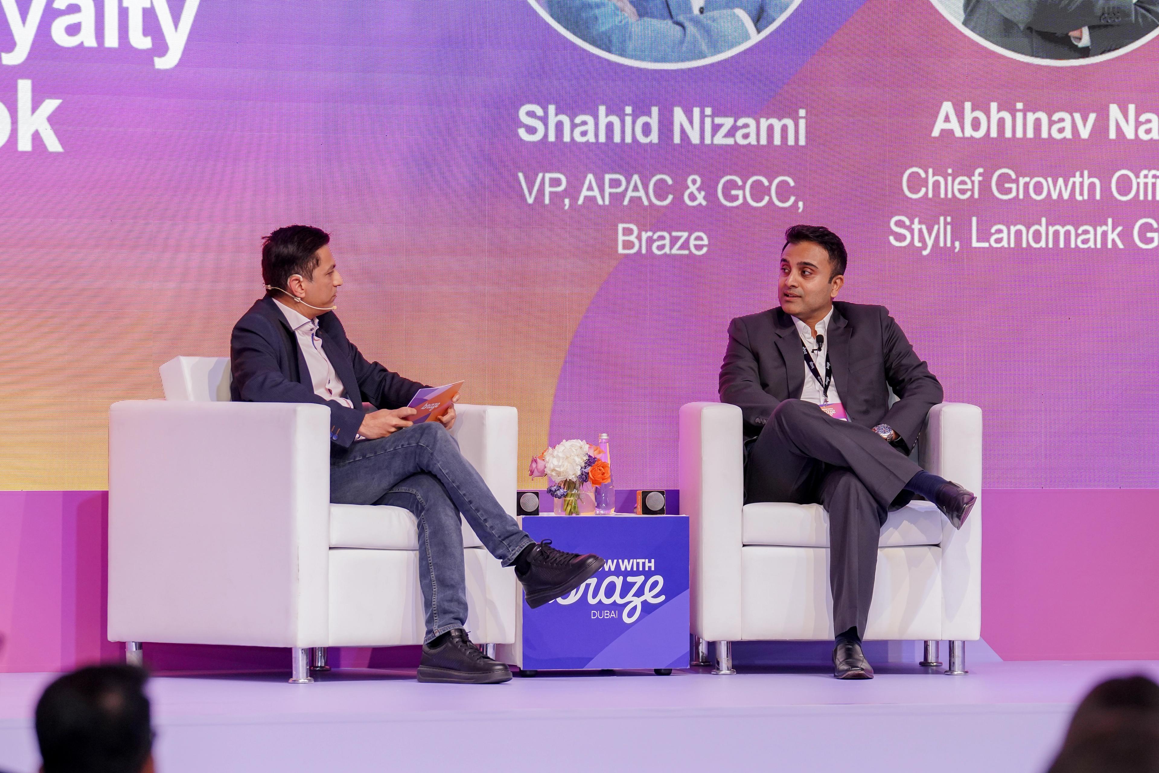 Two men seated on a stage, participating in a panel discussion with names and titles on the backdrop.