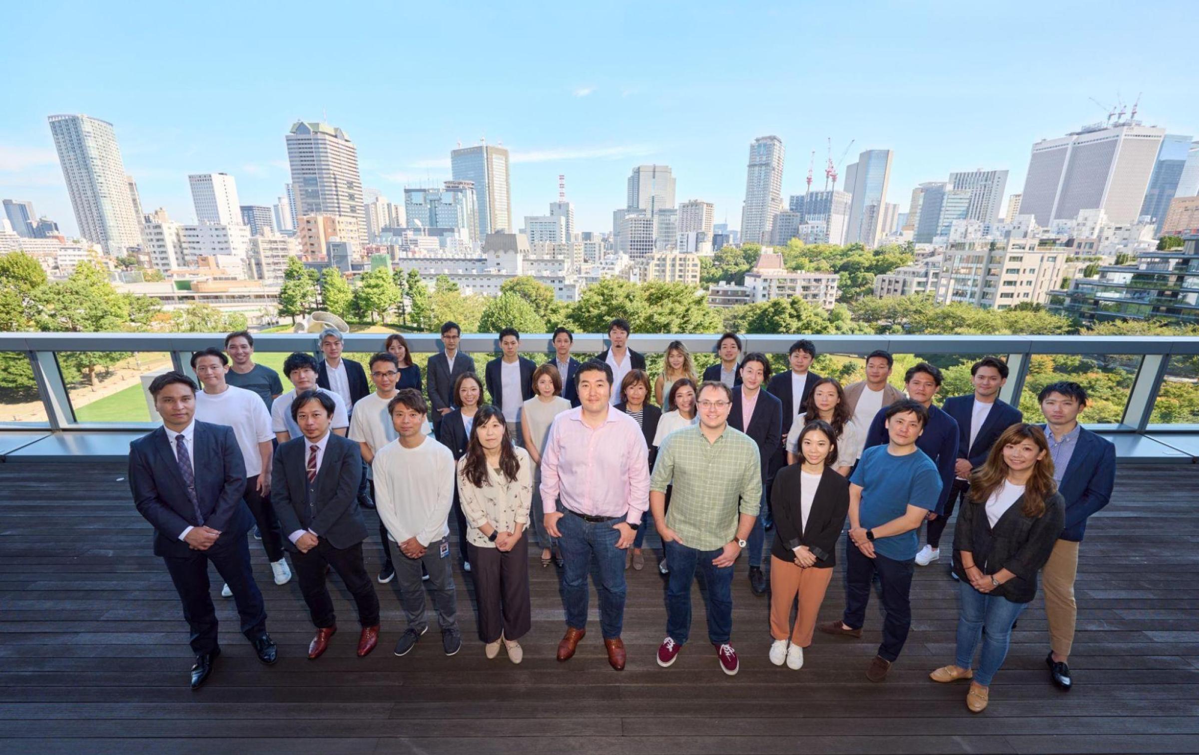 A diverse group of people stands on a balcony with a city skyline in the background. They are dressed in business casual attire and smiling for the photo.