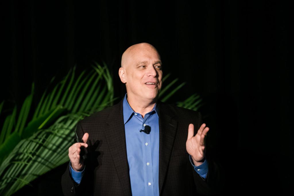 A man with a bald head and a blue shirt speaks on stage, gesturing with his hands. A green plant is visible in the background.