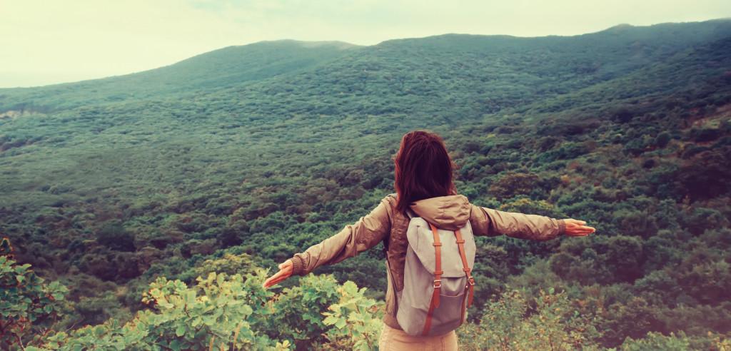 A person with a backpack stands with arms outstretched, overlooking a lush green landscape and mountains in the distance. The scene conveys a sense of freedom and connection with nature.
