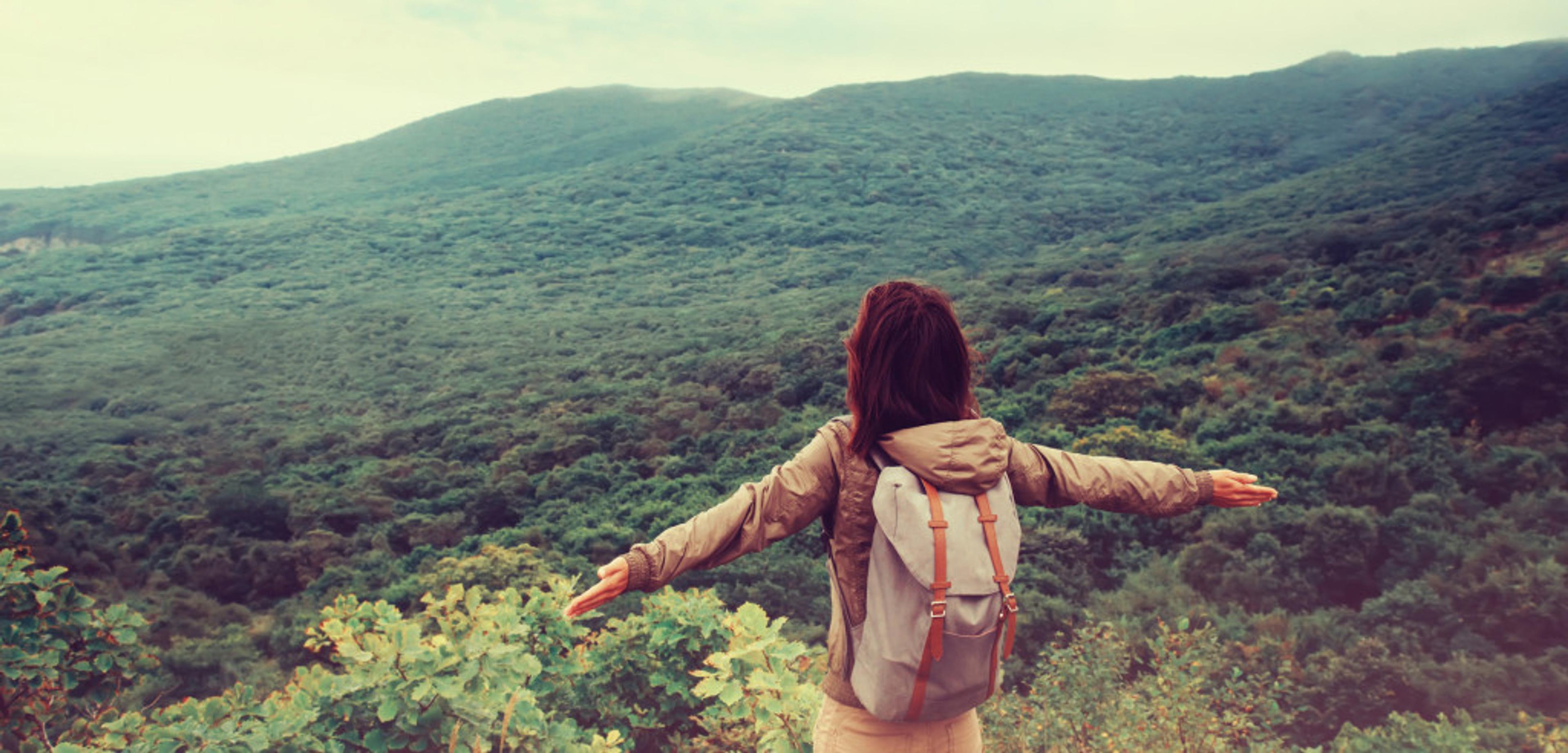 A person with a backpack stands with arms outstretched, overlooking a lush green landscape and mountains in the distance. The scene conveys a sense of freedom and connection with nature.
