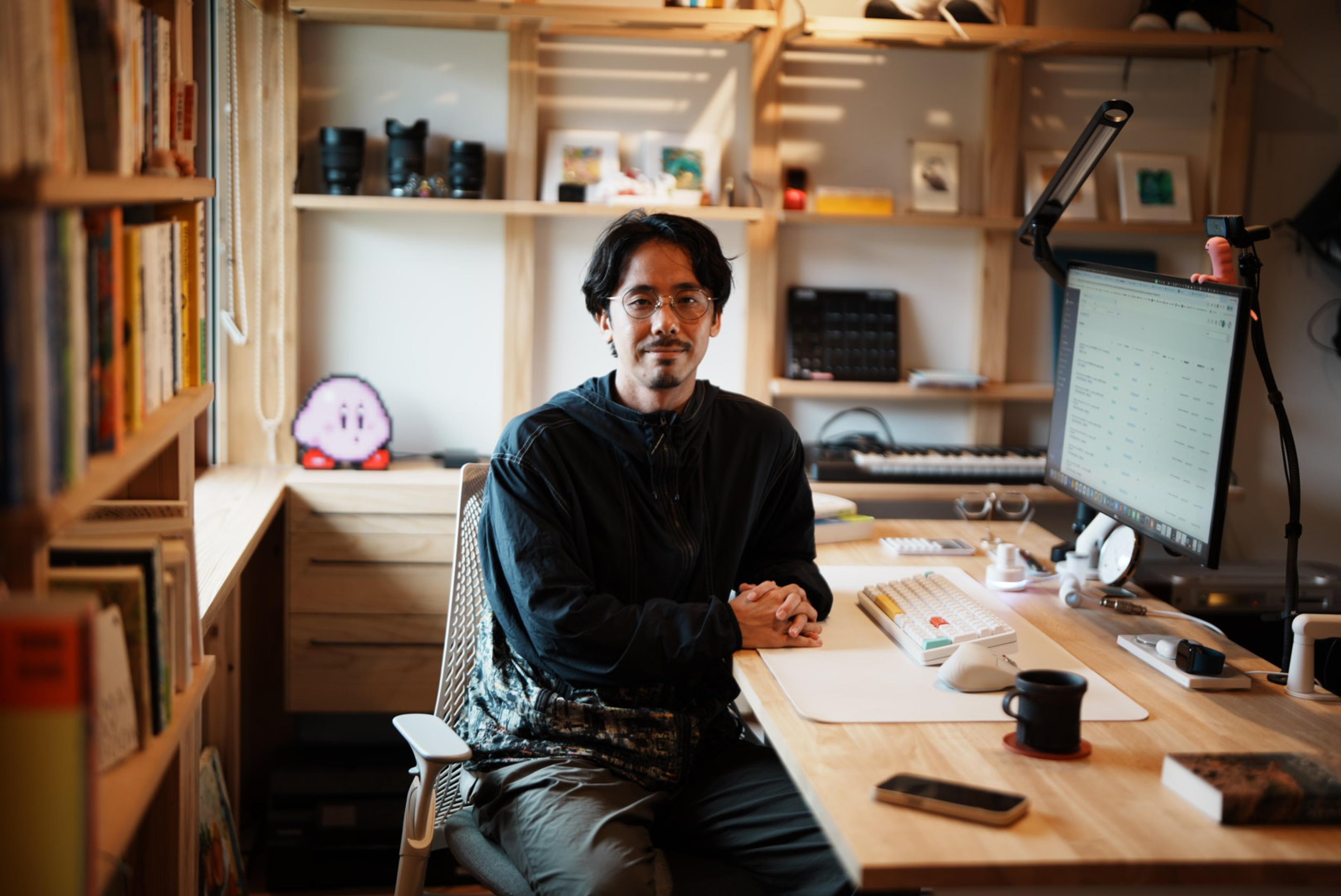 本棚や機材に囲まれた仕事部屋で、眼鏡をかけた男性がパソコンのある机に座っている。