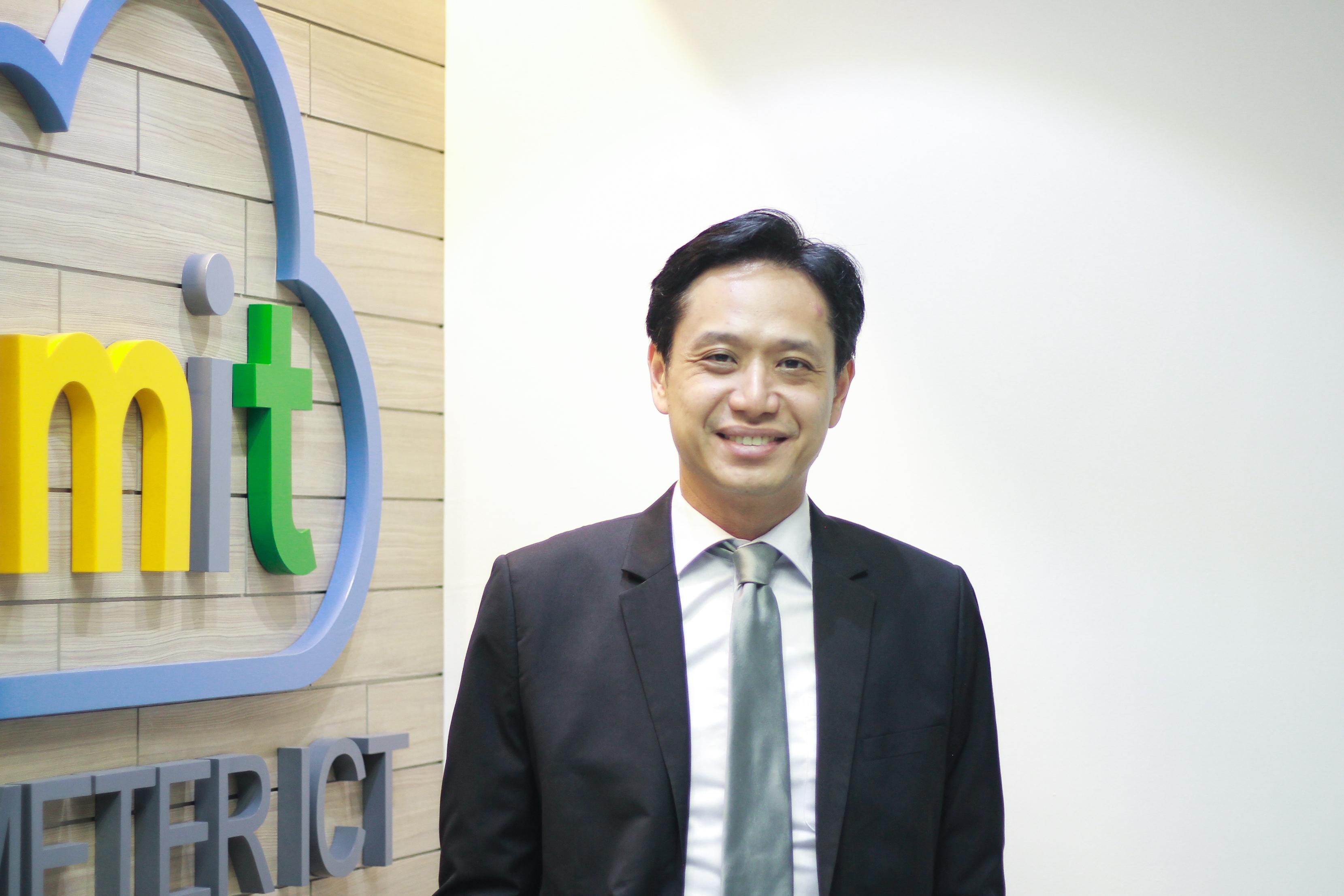 A man in a suit stands smiling in front of a colorful company logo on a wooden wall.