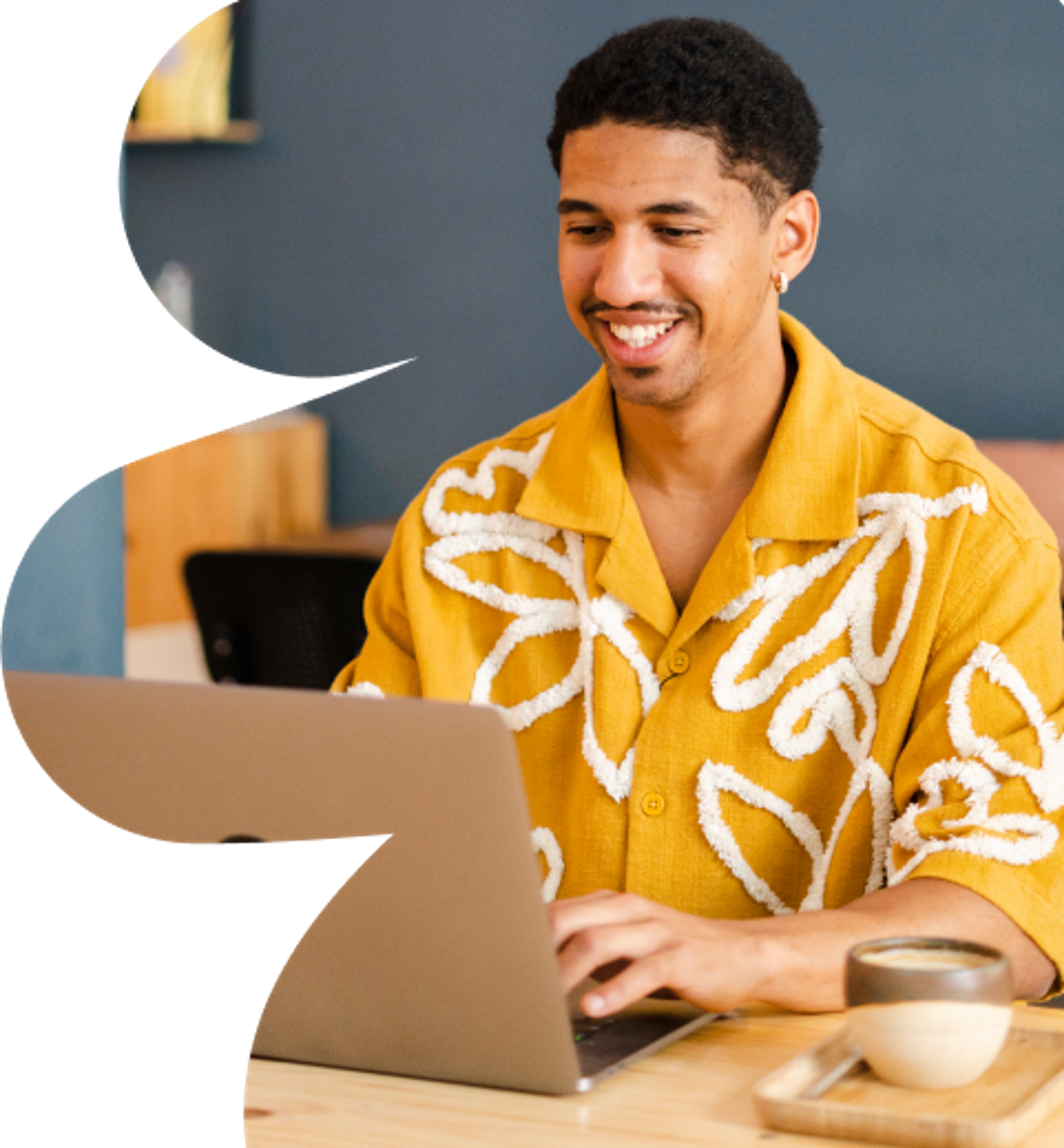 A man in a yellow shirt with white patterns smiles while working on a laptop at a table. A cup is placed in front of him.