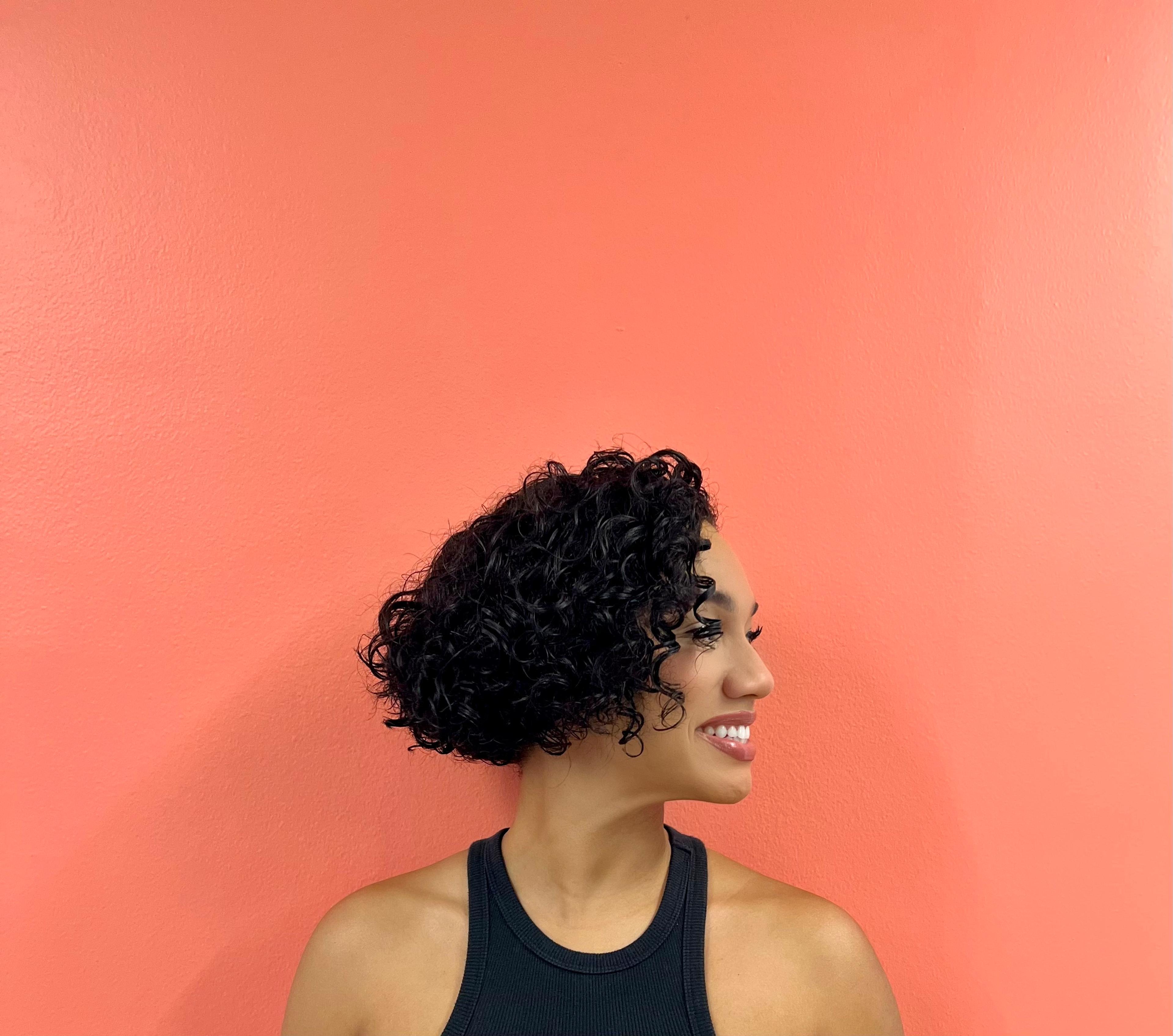 A woman with curly hair smiles while facing sideways against a coral-colored wall. She is wearing a black tank top.