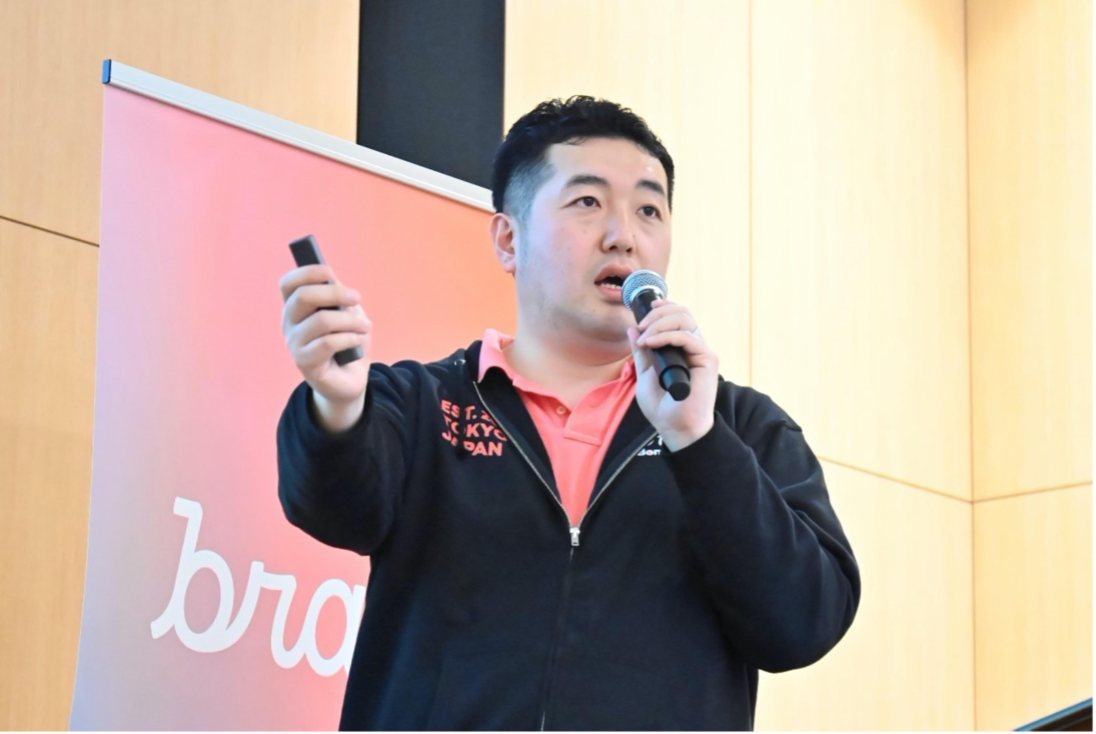 男性がマイクを持って話している様子。背景には「brand」のロゴが見える。