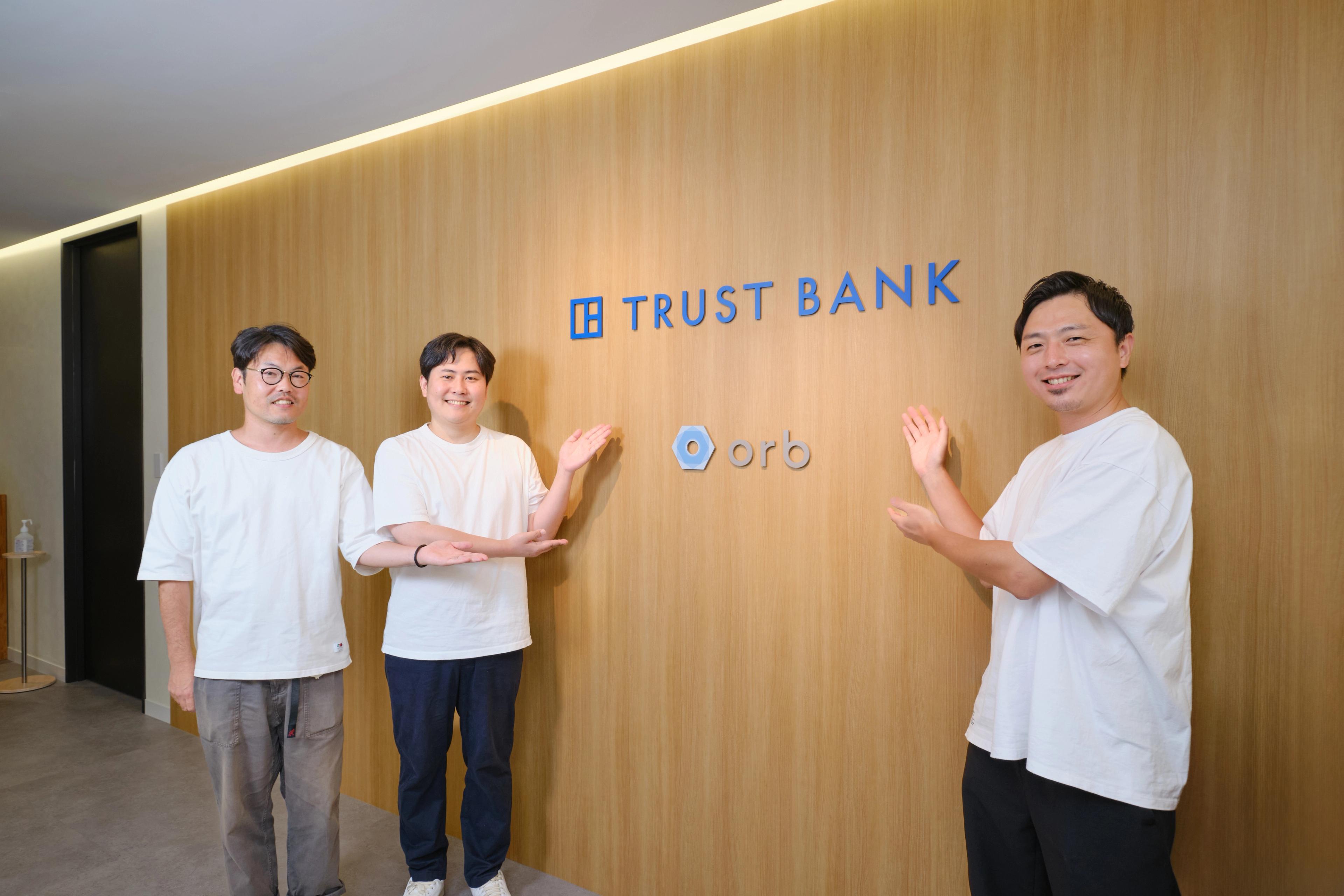 TRUST BANKのロゴの前でポーズをとる3人の男性。全員が白いTシャツを着ている。