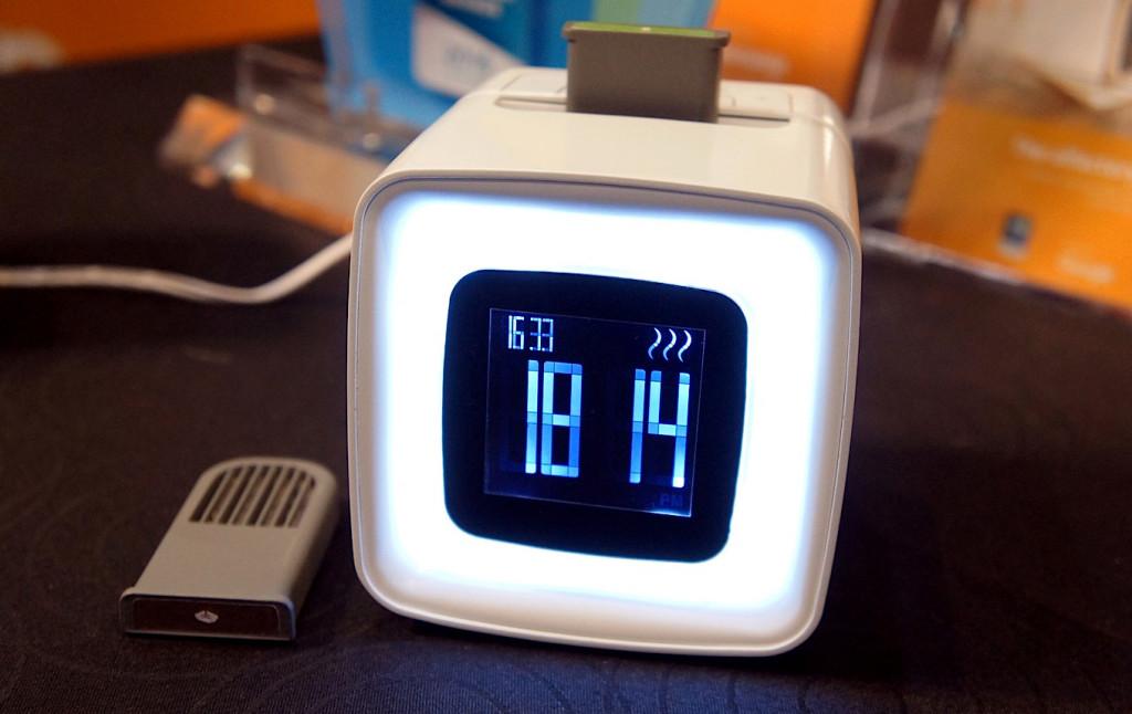 A modern digital alarm clock with a square design, displaying the time and temperature on a bright screen. It features a white body and a green top.