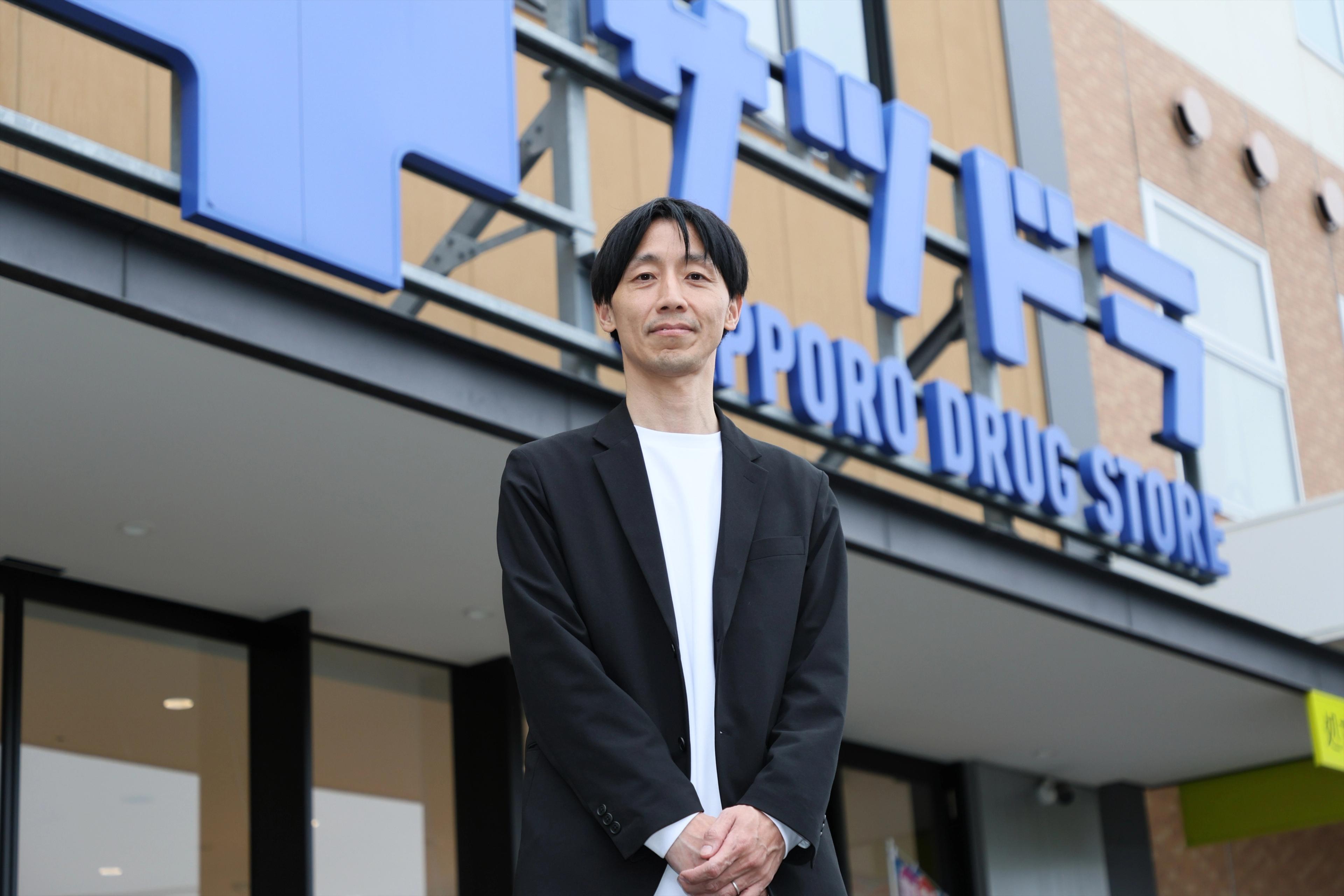 サツドラホールディングス株式会社 CDO兼株式会社サッポロドラッグストアー OMO本部本部長兼マーケティング部ゼネラルマネージャーの坂本 武史さんがサツドラロゴのビルの前でたっている
